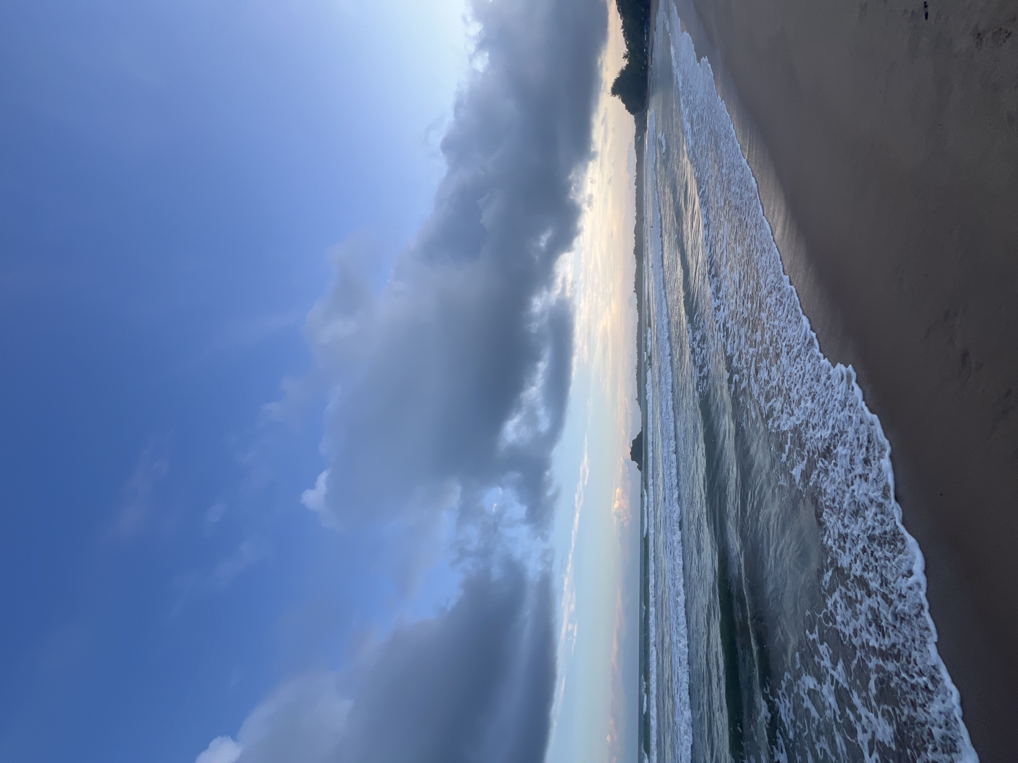 This is a photograph of a beach in Peleana, Sri Lanka, at dusk. The sky is a soft blue with scattered clouds, some of which are dark and heavy. The ocean is calm with gentle waves rolling onto the shore. The sand is wet and dark, reflecting the soft light of the setting sun. There are a few people in the distance, appearing as small figures on the beach and in the water, likely enjoying the evening. A rocky outcrop is visible on the horizon to the left, and a treeline marks the edge of the land to the right. The overall scene is peaceful and serene, capturing the beauty of a tropical coastline as day transitions to night.