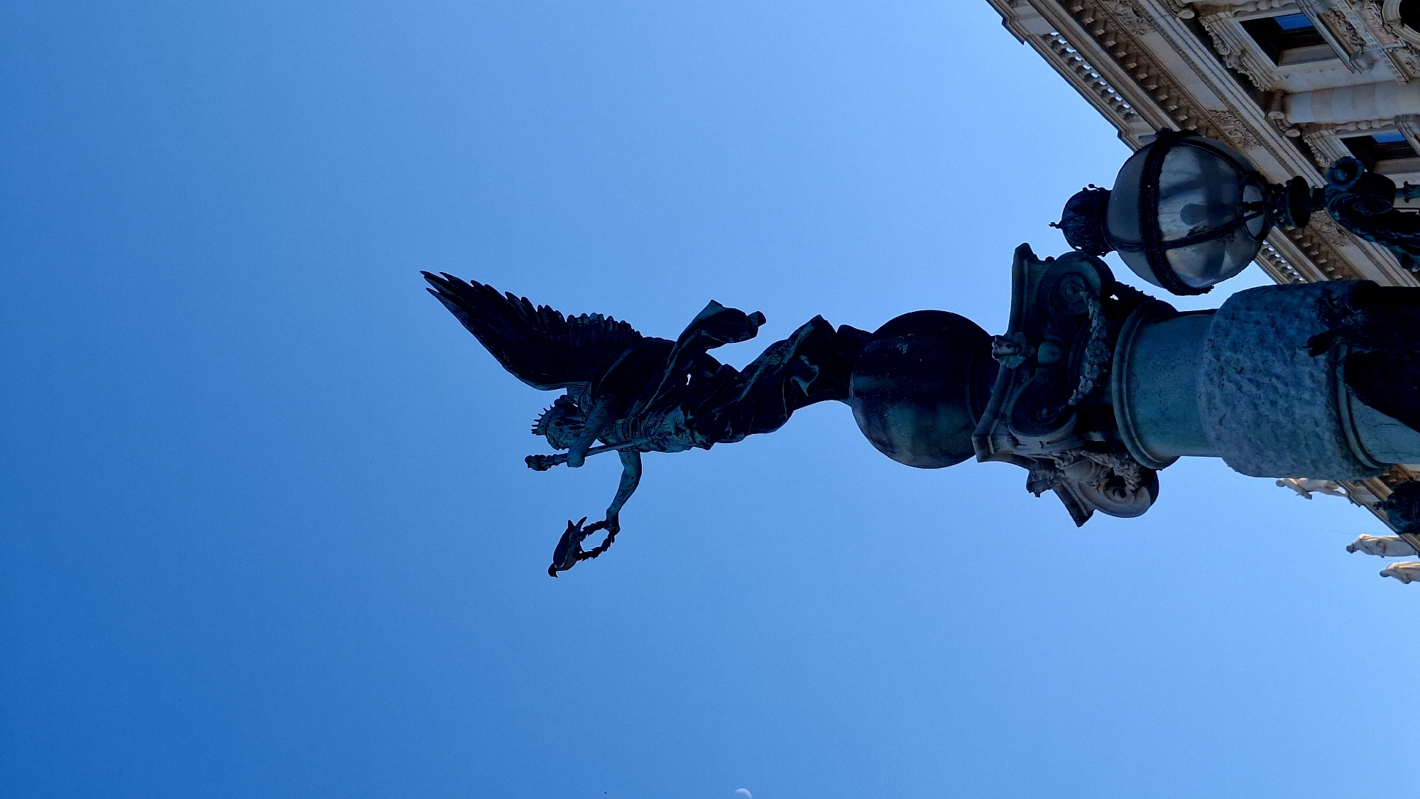 This is a low-angle, close-up, vertical shot of the Pallas Athene statue, located in front of the Austrian Parliament Building in Vienna, Austria. The statue, made of bronze, depicts Pallas Athene standing on a globe, holding a scepter in her right hand and a laurel wreath in her left. A bird is perched on the wreath. The statue's wings are spread, and its drapery flows. In the background, the sky is a clear, bright blue, with a sliver of the moon visible on the left. To the right of the statue, a portion of the ornate facade of the Parliament Building and a streetlamp are visible. The statue is dark, silhouetted against the bright sky, and appears weathered.