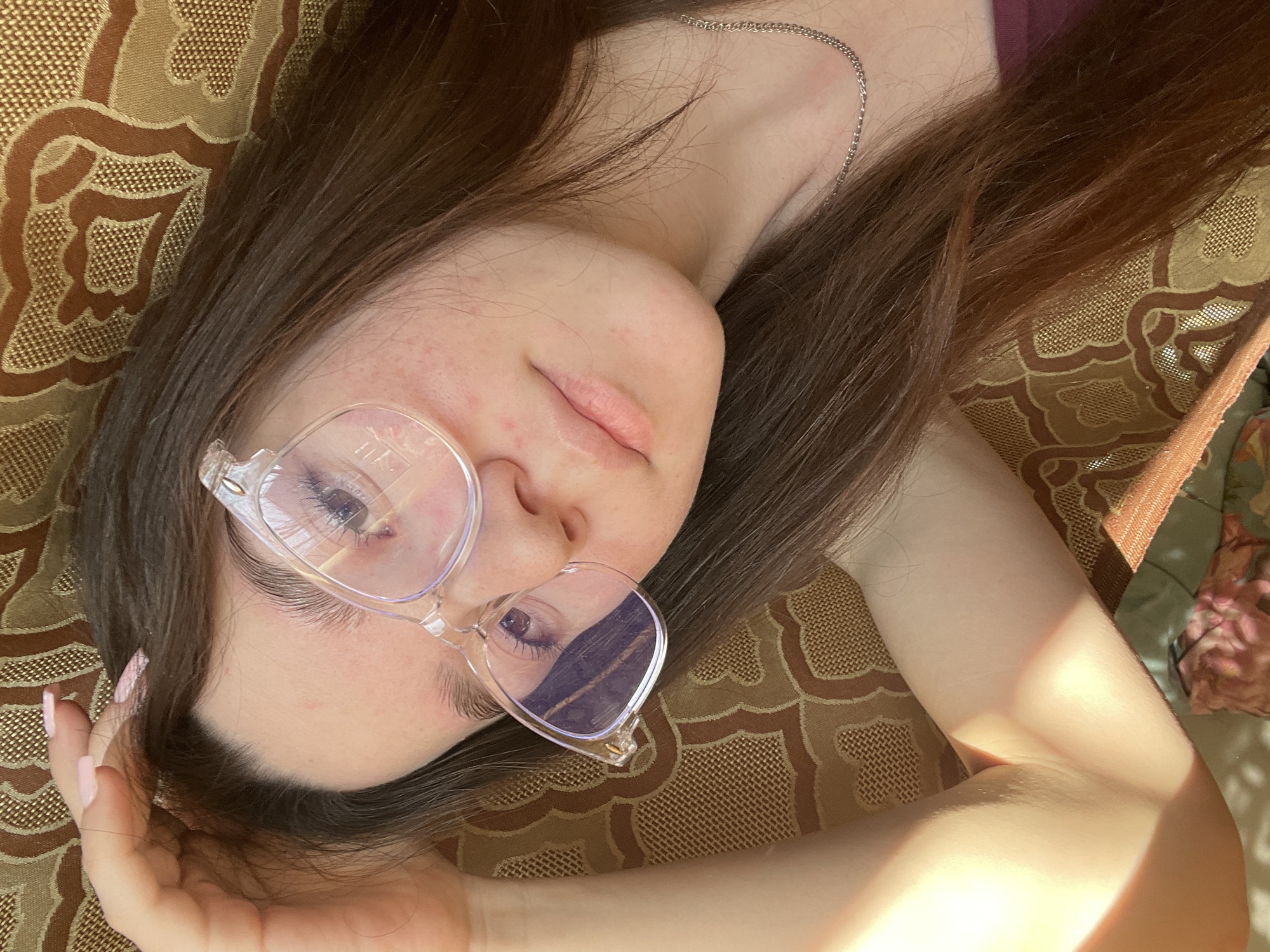 A young woman with long brown hair, wearing clear-framed glasses and a silver necklace, is lying on a patterned tan and brown surface. Her left arm is raised and partially obscures the right side of her face. The location is Ольховка, Russia.