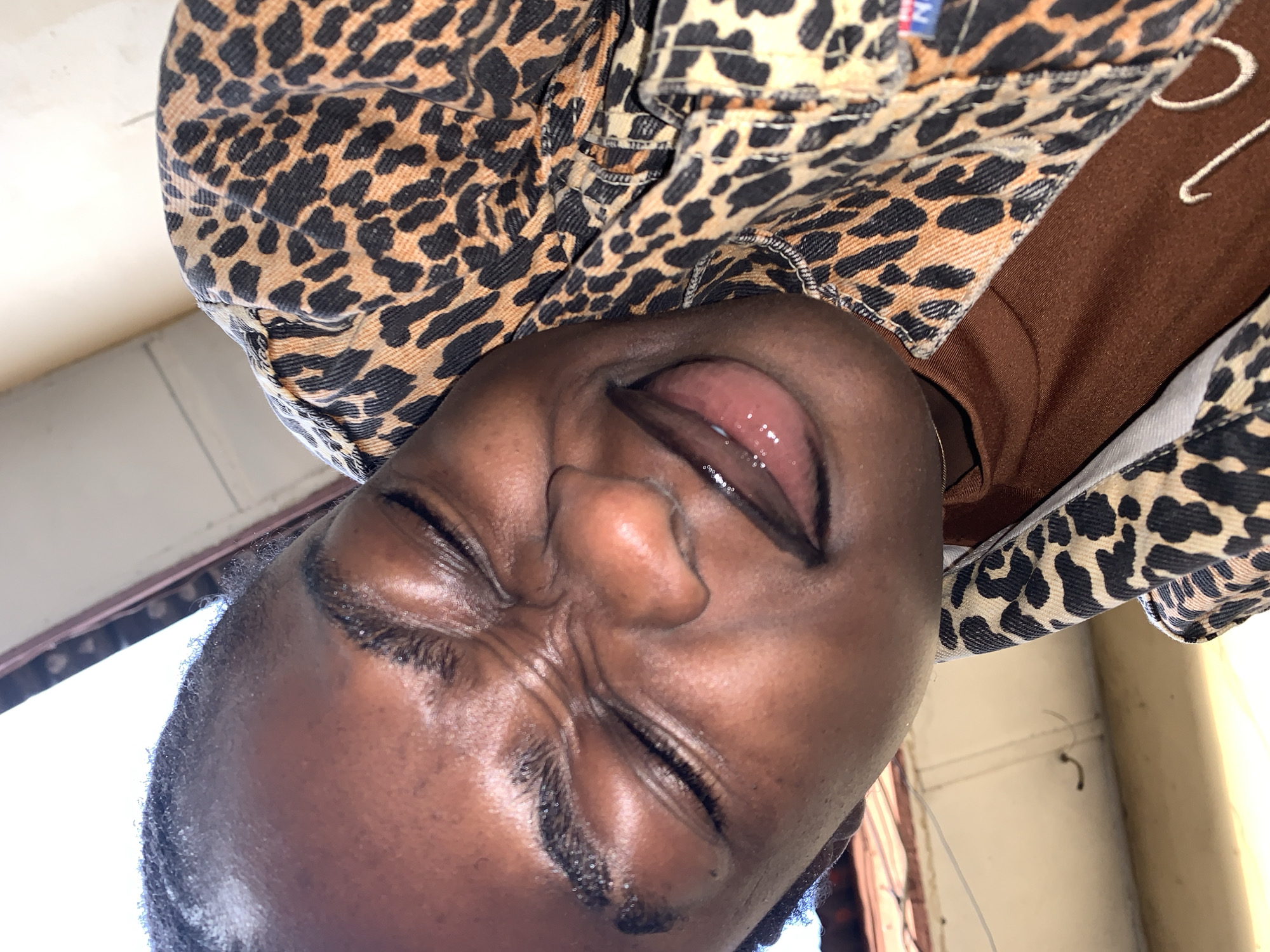 The image is a close-up, low-angle selfie of a young Black person with their eyes closed and face scrunched in an expression of discomfort or laughter. They are wearing a brown t-shirt under a leopard-print jacket. The background appears to be an indoor environment with a light-colored wall and ceiling, and a glimpse of what looks like outdoor elements through an opening. The lighting suggests it's daytime, with strong light coming from above and to the left. The overall impression is of a casual, personal moment captured in what might be a domestic setting. No specific location in Rafin Par, Nigeria can be definitively identified from the visual information provided.