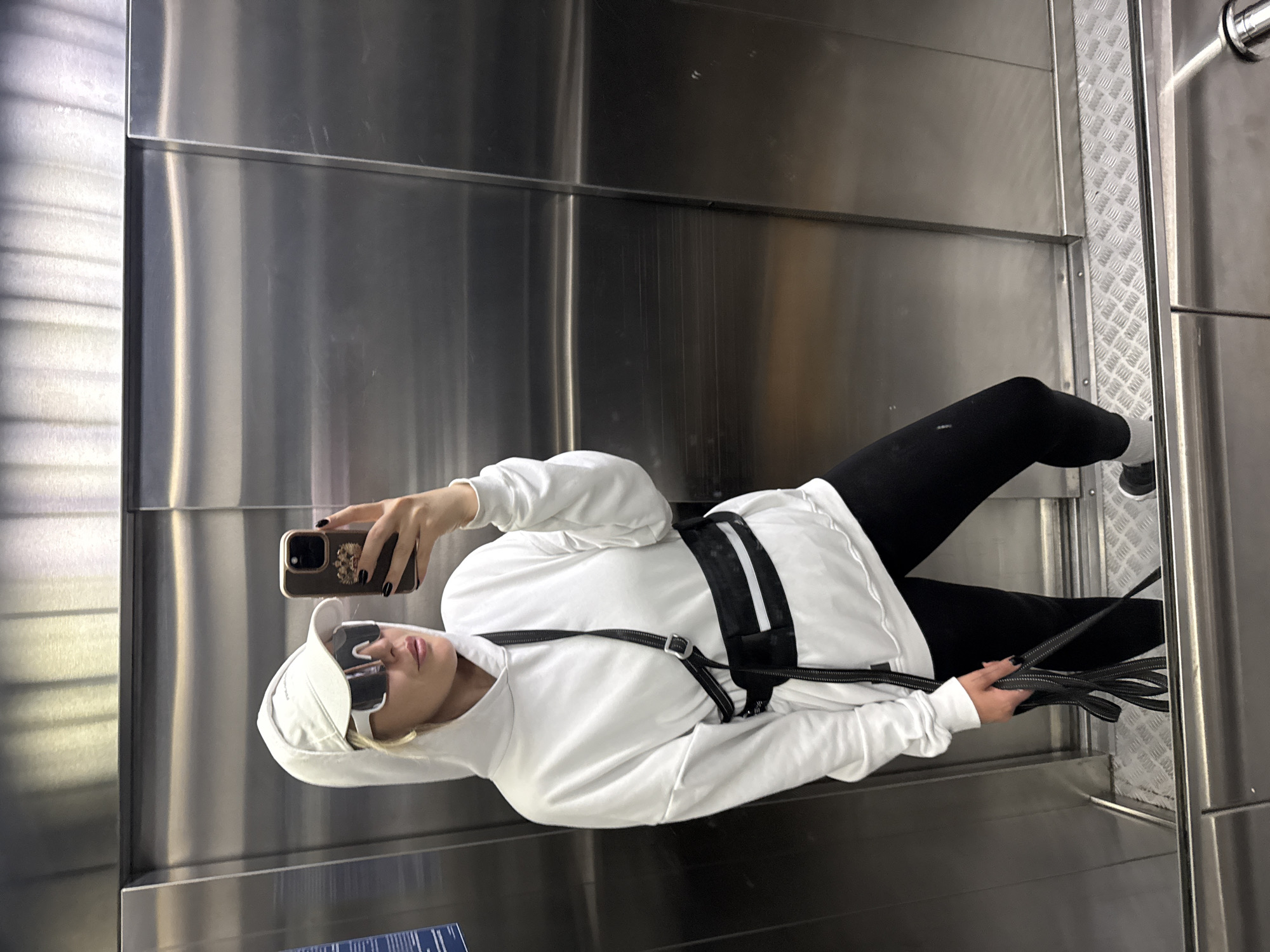 A person is lying on their side in what appears to be a commercial elevator. The elevator has stainless steel walls and a diamond-plated floor. The person is wearing a white hooded sweatshirt, black leggings, white socks, and sunglasses. They are holding a cell phone in their right hand, taking a selfie. Their left hand is holding a collection of black cables. They have a black harness around their torso. The location is presumed to be a building in Krasnoyarsk, Russia.