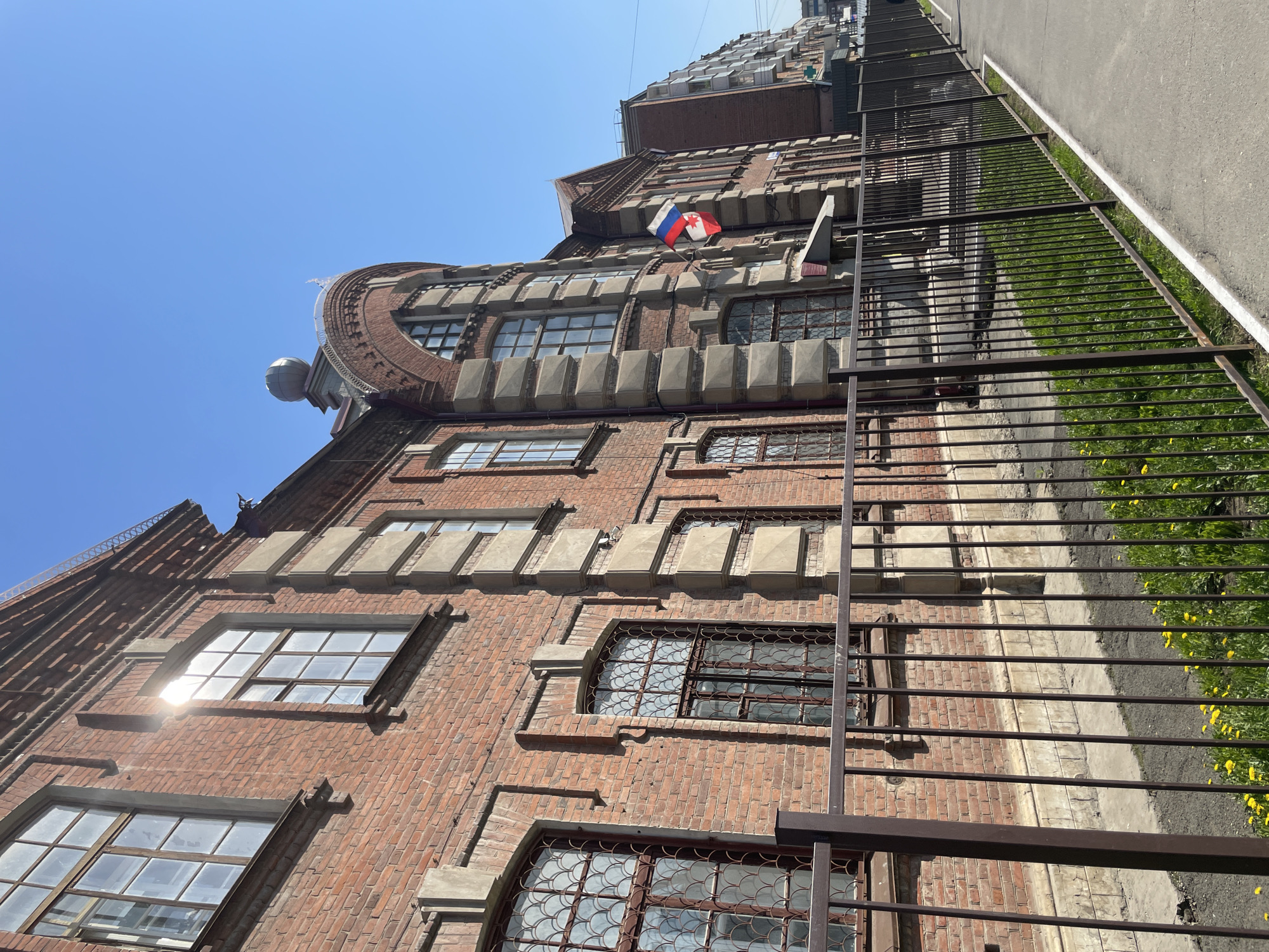 The image shows a brick building in Izhevsk, Russia, viewed from a low angle. The building has multiple windows, some with decorative metal grilles. A Russian flag is visible on the upper portion of the building. A metal fence runs along the side of the building, bordering a paved area with some vegetation visible. The sky is clear and blue.
