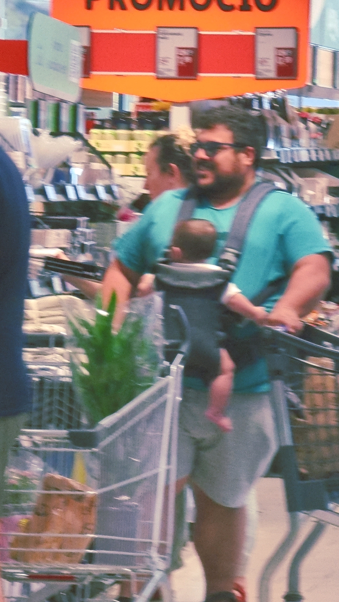 The image shows a man wearing sunglasses and a teal t-shirt, carrying a baby in a carrier. The man is pushing a shopping cart. A portion of a woman wearing a teal t-shirt is visible on the left side of the image. In the background, there are shelves stocked with various items. A promotional sign with orange and red colors is visible at the top of the image. The scene appears to be in a supermarket. The lighting suggests it is daytime. There are no specific landmarks visible to determine the location in Olot, Spain.