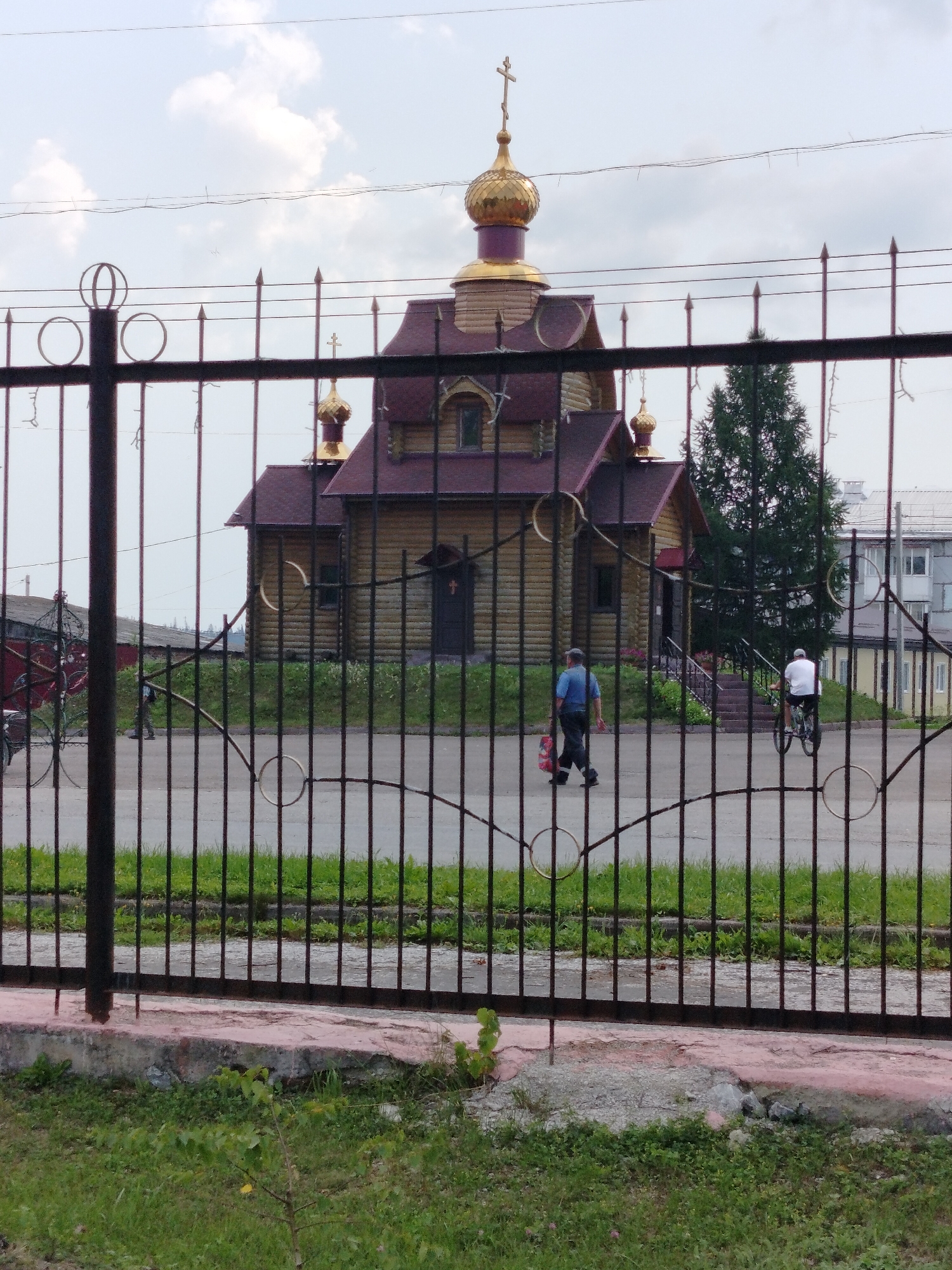 The image depicts a wooden church with golden domes, situated in Yashkino, Russia. In the foreground, a black metal fence with decorative circular elements obstructs the view of the church and its surroundings. Behind the fence, a man in a blue shirt and dark pants is walking away from the camera, carrying a red bag. To his right, a person on a bicycle wearing a white shirt and dark shorts is riding away from the camera. The church has a dark red roof and is made of logs. A tall green tree stands to the right of the church. The sky is partly cloudy. The ground in front of the fence is covered in grass, and there is a paved area behind the fence.