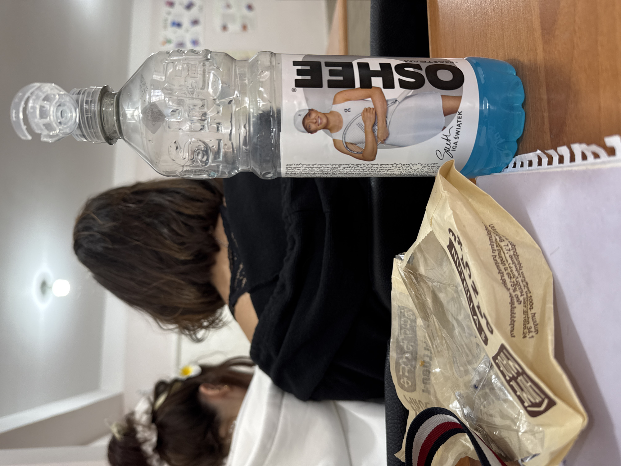 The image shows a partially filled, transparent plastic water bottle labeled "OSHEE" with an image of Iga Świątek on it, resting on a desk with a notebook. Next to the bottle is a brown, crinkled paper bag labeled "NUTELLA". A person with dark hair is seated at the desk, partially visible in the background. Another person is visible in the lower-left corner of the frame. The background is out of focus, but a white wall and lighting fixtures are visible. The image includes partial OCR text in Armenian. The location of the image may be Yerevan, Armenia based on the OCR in Armenian.