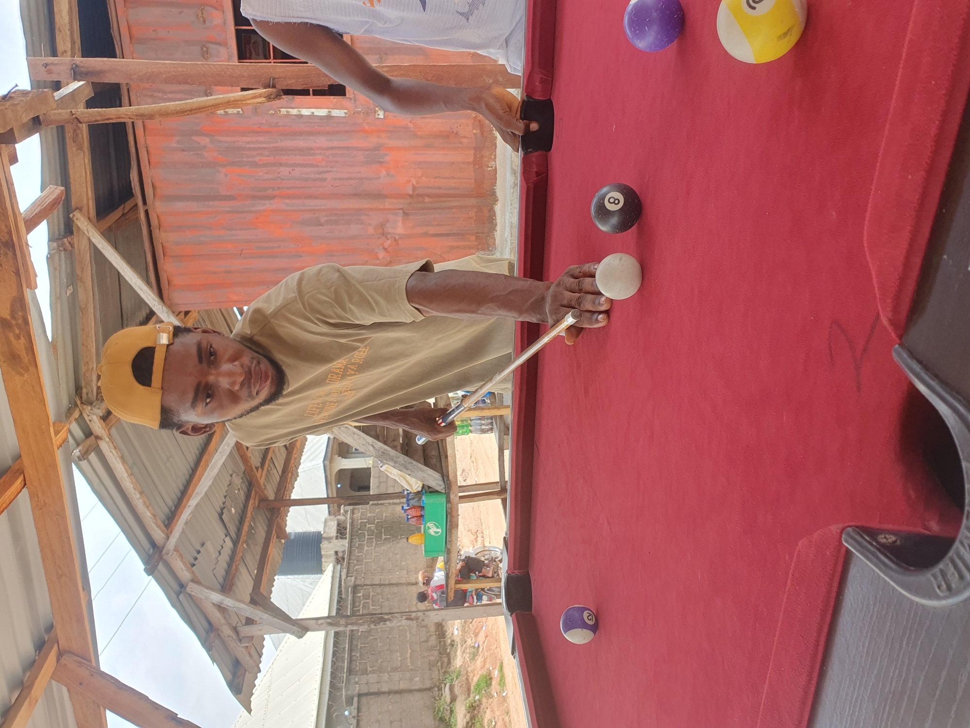 In Lokoja, Nigeria, a man in a yellow cap and a tan shirt is playing pool. He is aiming his cue stick at a white ball. Several other pool balls are on the red felt-covered table, including a purple ball with the number 2, a black ball with the number 8, a purple ball with no visible number, and a yellow and white striped ball. The pool table is located outdoors under a corrugated metal roof supported by wooden beams. In the background, a metal wall with orange corrugated siding can be seen, along with a brick wall and some buildings. Two people are visible in the background, one on a motorcycle and another sitting nearby, behind a green table with bottles on it.