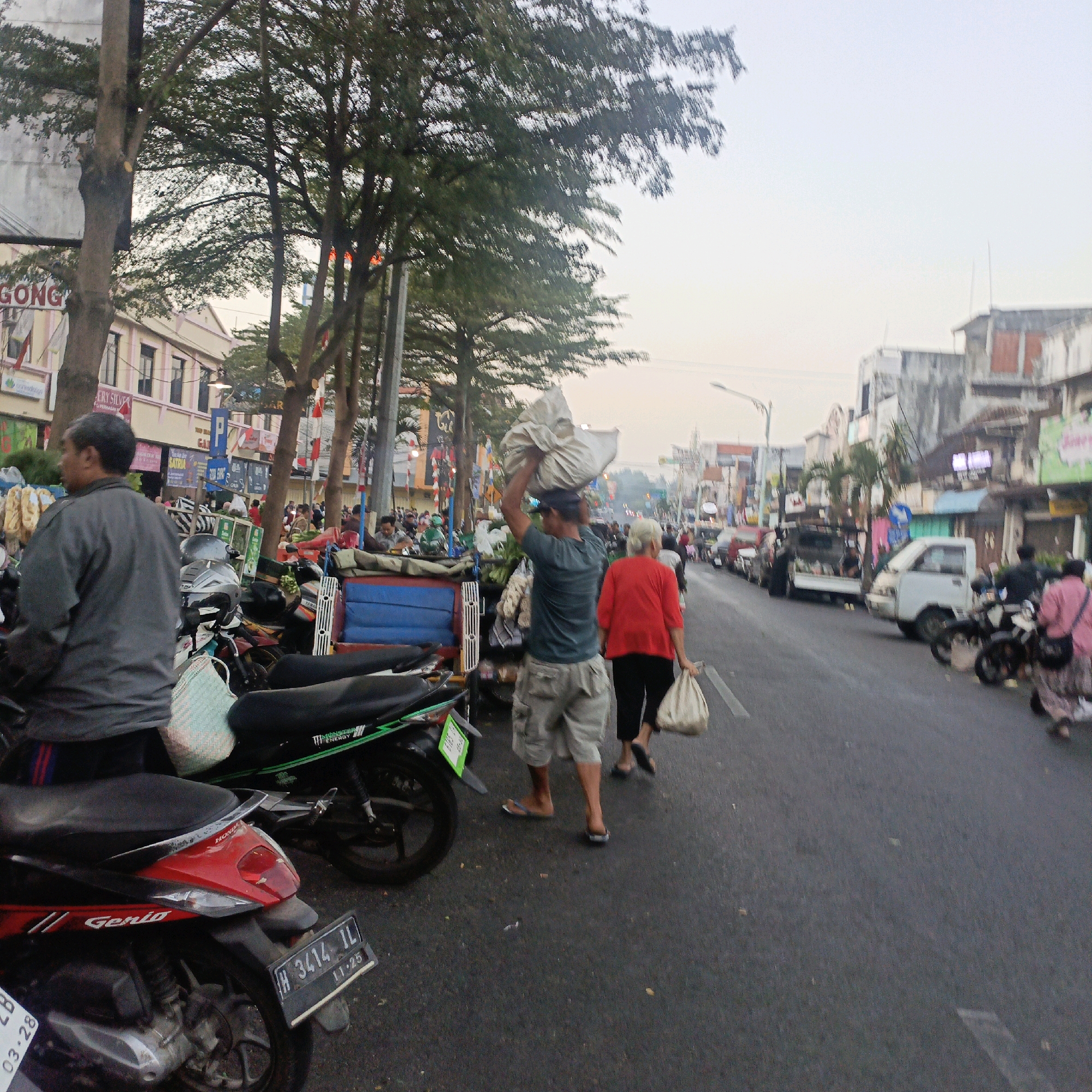 The image depicts a bustling street scene, likely in Salatiga, Indonesia, with several people engaged in activities. In the foreground, a man in a grey shirt is seen from the side, facing left, with motorcycles parked behind him. One motorcycle in the immediate foreground is red and black, displaying a license plate "H 3414 IL" and the word "GENIO". Another motorcycle to its left is black with green accents, bearing the text "MONSTER ENERGY".
Further into the scene, a man is carrying a large white sack on his head while walking away from the camera. He is wearing a dark t-shirt and light-colored cargo shorts. A woman in a red shirt and black pants is walking ahead of him, also carrying a white bag in her right hand. A second woman in a pink and white patterned outfit is seen to the far right, also with a white bag.
The background reveals a street lined with buildings and trees. Some of the buildings appear to be shops or market stalls, with visible signage. The street is paved and appears to be a public thoroughfare with traffic, including a white van and several motorcycles. The overall atmosphere suggests a busy market or commercial area, possibly during the late afternoon or early evening given the lighting.
