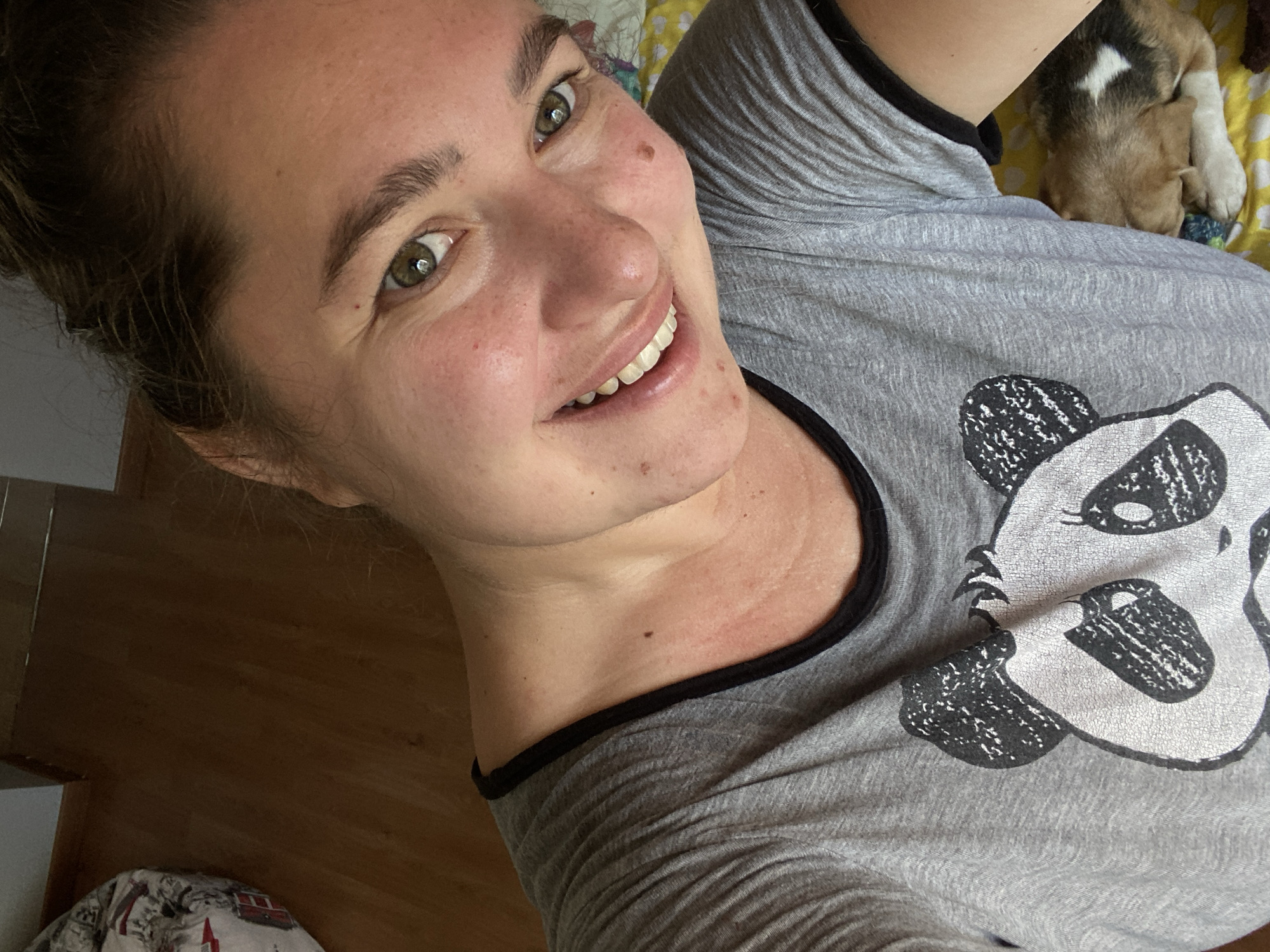 The image shows a close-up, selfie-style portrait of a young woman with brown hair pulled back, green eyes, and a wide smile. She is wearing a grey t-shirt with a black panda graphic on it. A small, tri-colored puppy is visible sleeping near her right shoulder. The background consists of a wooden floor and a patterned blanket. The image does not provide any information that would allow for the determination of its location within the city of Perm, Russia, or any other specific geographic location.