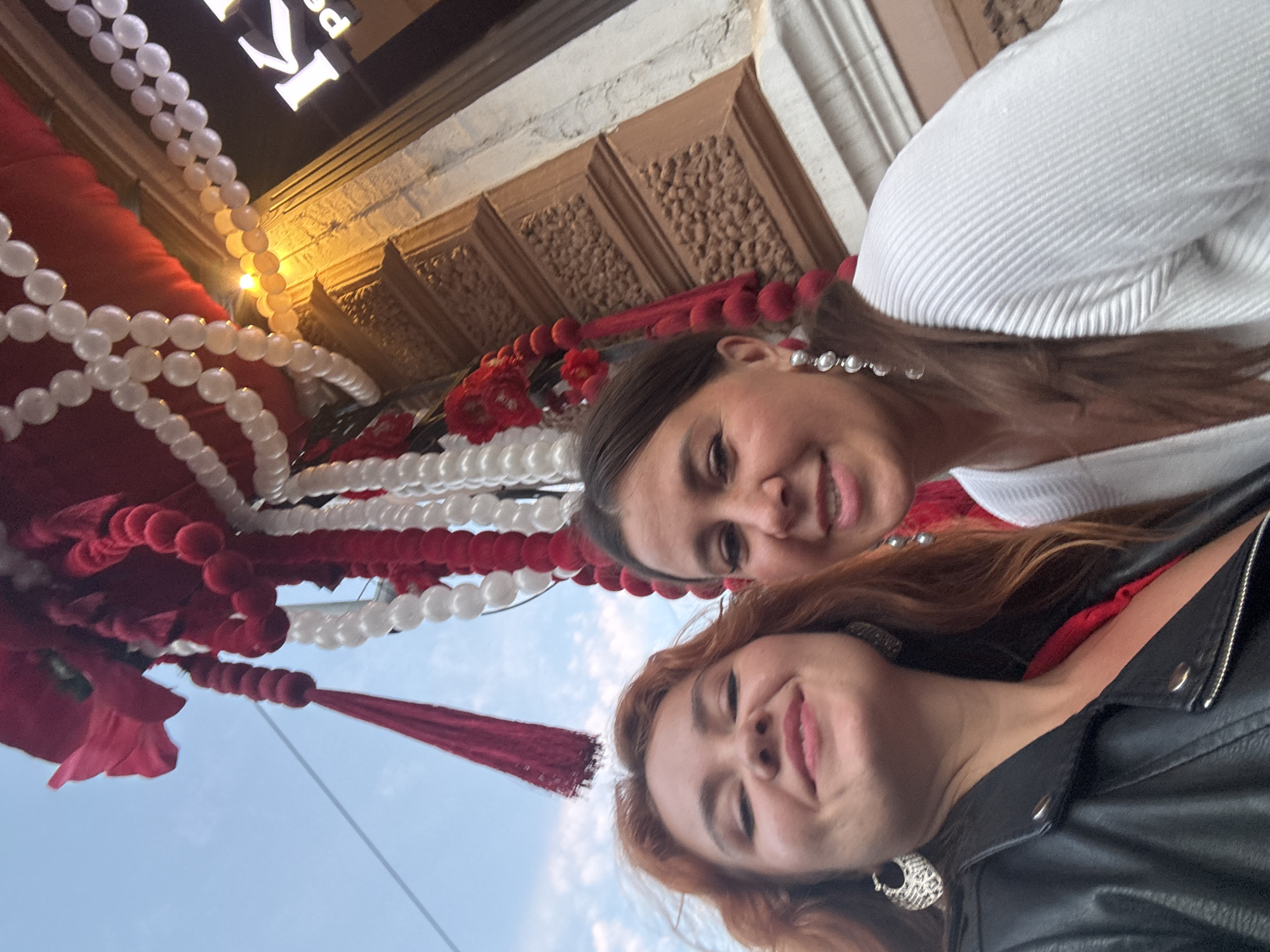 Two women are taking a selfie outdoors in front of a building with ornate brown architectural details. They are positioned in the lower half of the frame. The woman on the left has long, auburn hair and is wearing a black leather jacket and large, silver hoop earrings. She is smiling directly at the camera. The woman on the right has brown hair and is wearing a white, ribbed sweater and long, dangling pearl earrings. She is also smiling. Above them, there is festive red and white balloon and fabric decoration hanging, possibly for a celebration or event. Part of a building facade with text that appears to be in Latin script is visible to the right. The sky above is a pale blue with some clouds. The overall lighting suggests it is either early morning or late afternoon. There is no explicit indication of the city of Nizhny Novgorod, Russia in the image.
