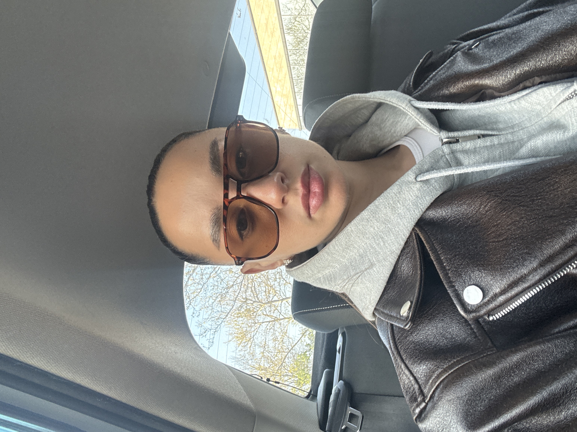 A person is depicted in a selfie-style shot from a low angle, leaning back in a car seat. The image is rotated counter-clockwise, making the person's head appear on the left side of the frame and their body extending to the right. The individual wears large, brown tortoise-shell framed sunglasses that obscure their eyes, and their lips are slightly parted. They have well-defined eyebrows and a neutral expression.
The person is dressed in a dark brown or black leather jacket, worn over a light grey hooded sweatshirt, with a sliver of a white garment visible beneath the hoodie. Details of the jacket, such as silver buttons and zippers, are visible. The car's interior is visible, featuring a grey fabric headliner and dark grey upholstery on the seats. A seatbelt buckle is noticeable on the right.
Through the car window, likely the rear or a side window, a bright outdoor scene is visible. Bare tree branches against a clear blue sky suggest a late winter or early spring day. Part of a modern building with a light-colored, possibly glass, facade can be seen in the upper background through the window. The overall lighting is bright, indicating daytime. No text is visible in the image. The location context is Belorechensk, Russia.