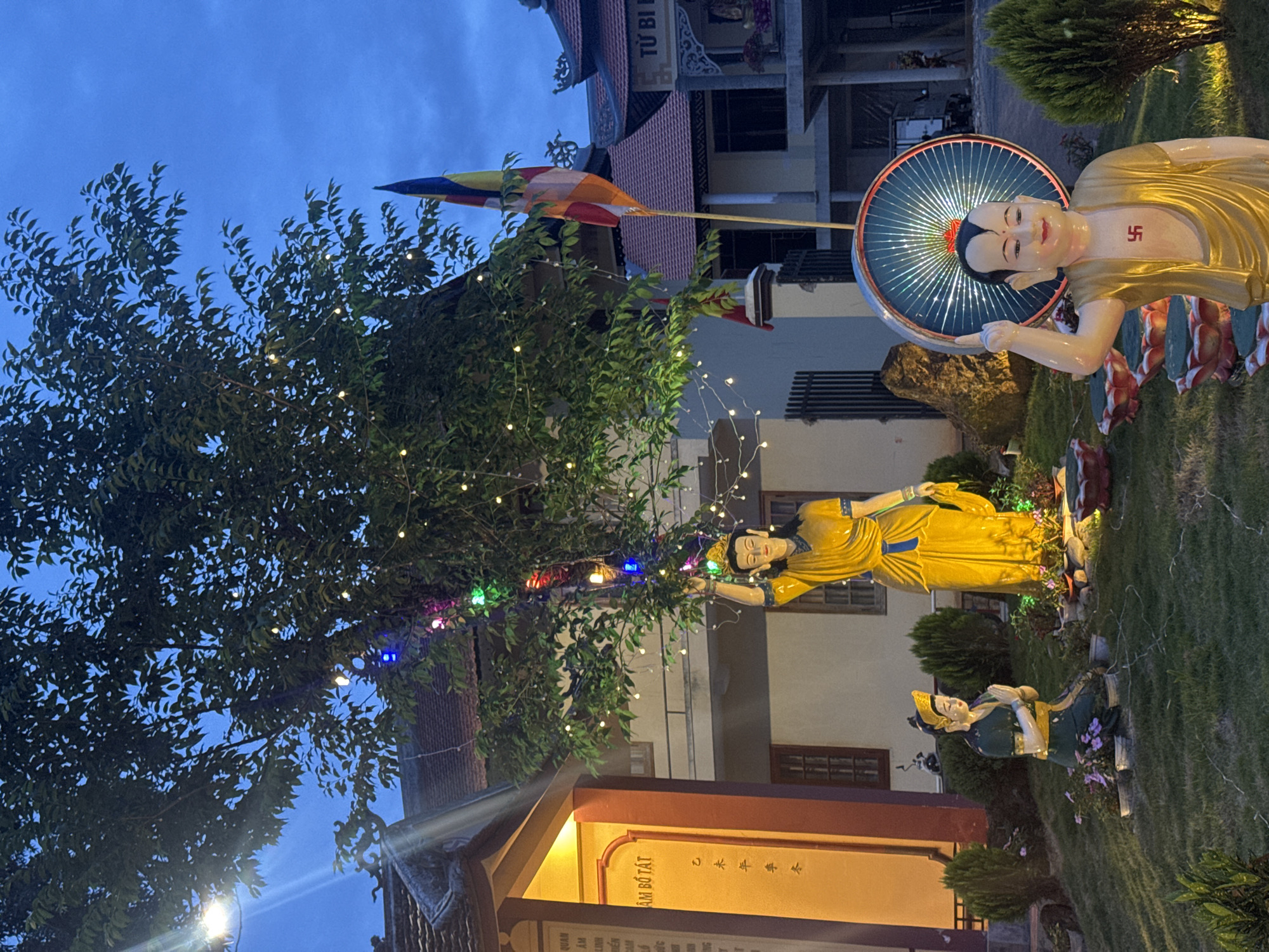 The image shows a temple scene in Da Lat, Vietnam, taken at dusk. A statue of a Buddhist deity with a halo stands prominently on the right, adorned with a swastika symbol on its chest. Another statue of a woman in a yellow robe is positioned near a tree decorated with small, colorful lights. A kneeling figure in green and gold attire is also present. A Buddhist flag flies in the background, partially obscured by the tree. The temple building is visible with signage in Vietnamese characters, illuminated by warm light. The environment is a well-maintained garden with neatly trimmed grass and shrubs. The sky is a deep blue.