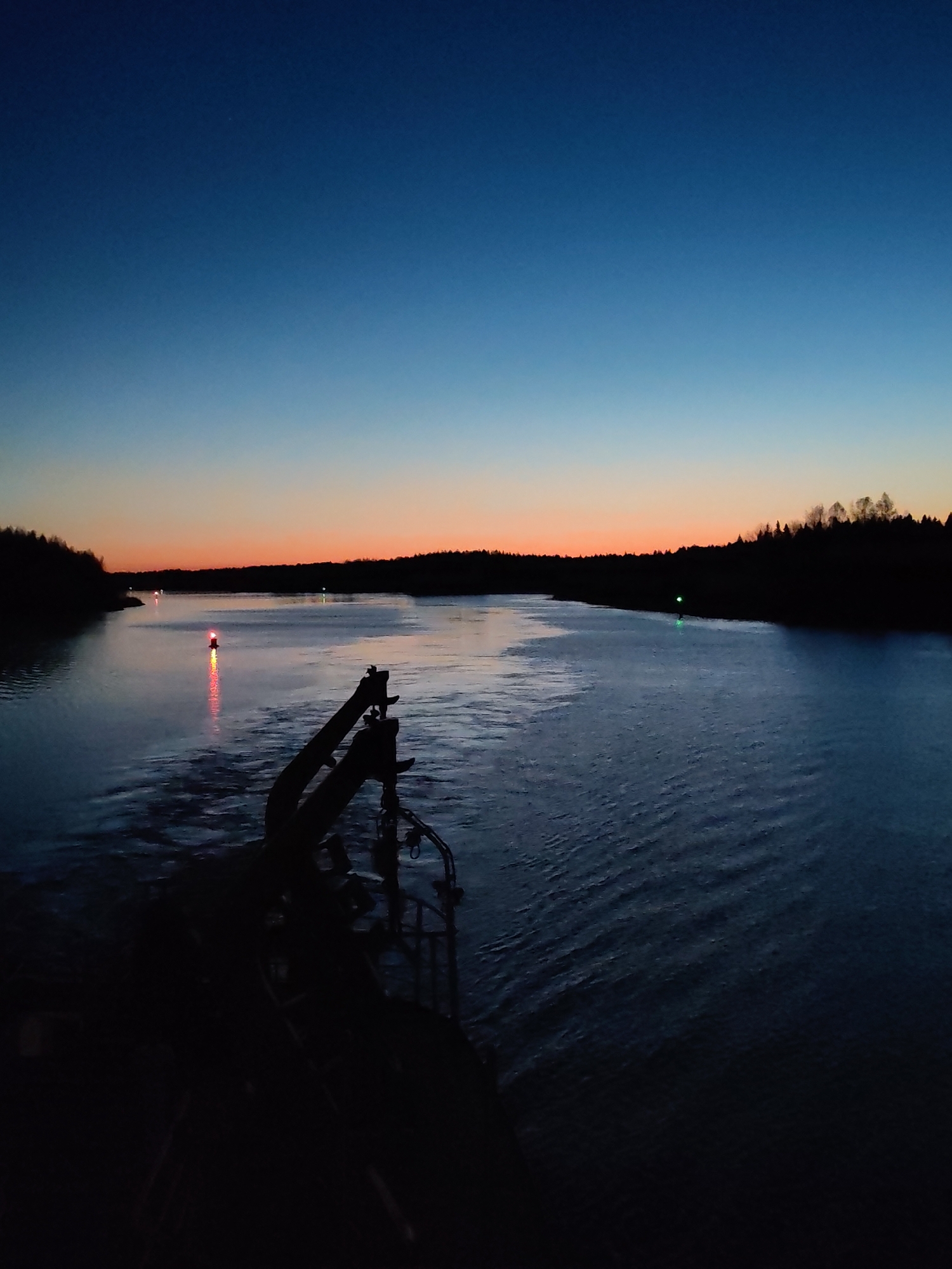 The image depicts a waterway at dusk, likely a river or a wide canal, in Badozhskoi Pogost, Russia. The foreground shows the dark silhouette of what appears to be the deck and crane of a vessel. The water is dark and rippled, reflecting faint light. Red and green navigation lights are visible on the water's surface, marking the channel. On either side of the waterway, the treeline is silhouetted against the horizon. The sky transitions from a deep blue at the top to orange and pink hues near the horizon, indicating either sunset or sunrise. There are no people visible in the scene. The focus is on the machinery of the vessel and the landscape.