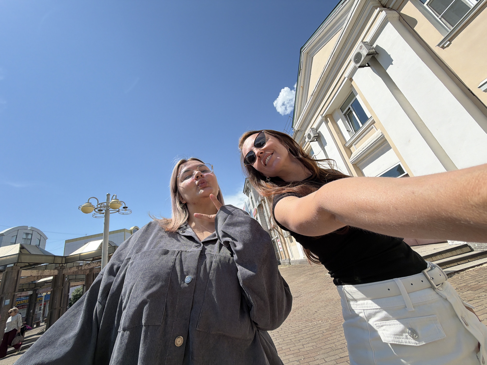 Two young women are posing for a selfie outdoors on a sunny day in Birobidzhan, Russia. The woman on the left is wearing glasses and a grey corduroy shirt, and she is making a duck face and holding up two fingers. The woman on the right is wearing sunglasses and a black top with a white skirt, and she is smiling. Behind them, a pale yellow building with white trim and windows can be seen, along with a blue sky with a single cloud. In the background, a street lamp and some architectural structures are visible. The ground is paved with bricks.