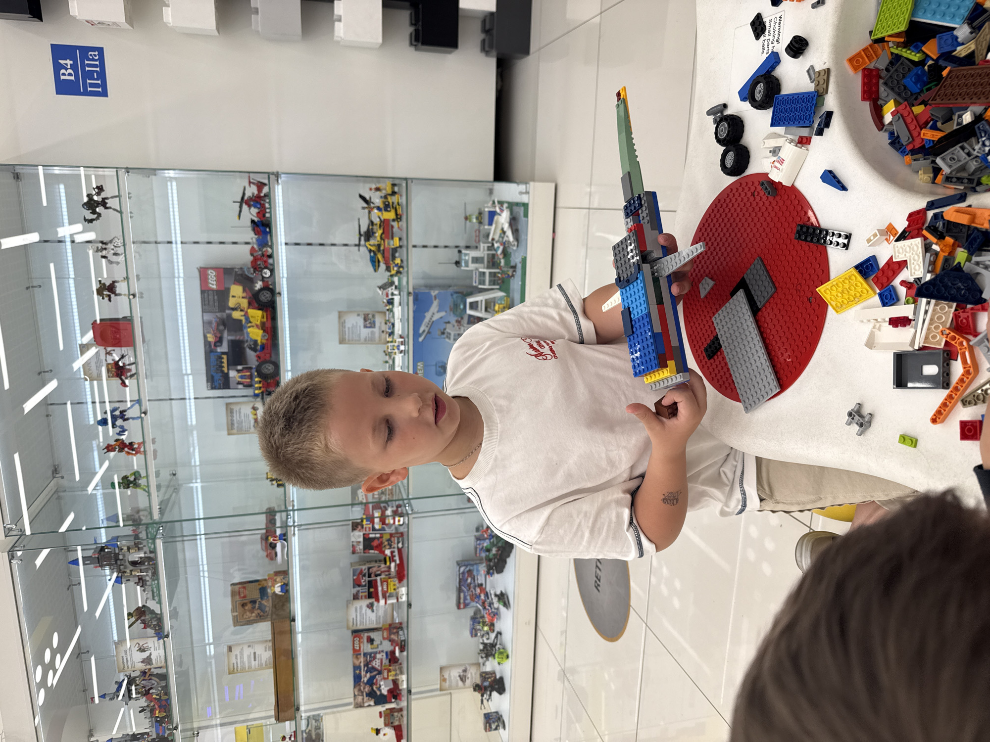 The image depicts a young boy in a white t-shirt and beige shorts constructing a Lego model, likely a spaceship, in a retail store environment. The store appears to be a Lego store or a toy store with a dedicated Lego section, as evidenced by the display cases filled with various Lego sets and models in the background. The boy is holding the partially assembled Lego creation in his hands, focusing on the construction. On the table in front of him are scattered Lego bricks, a red Lego baseplate, and other building components, indicating an ongoing activity. The store's flooring is tiled and white. A sign with Russian text "B4 П-Па" is visible on a white wall to the right. The overall scene suggests a child engaged in play and creativity within a commercial setting. This scene is located in the city of Khimki, Russia.