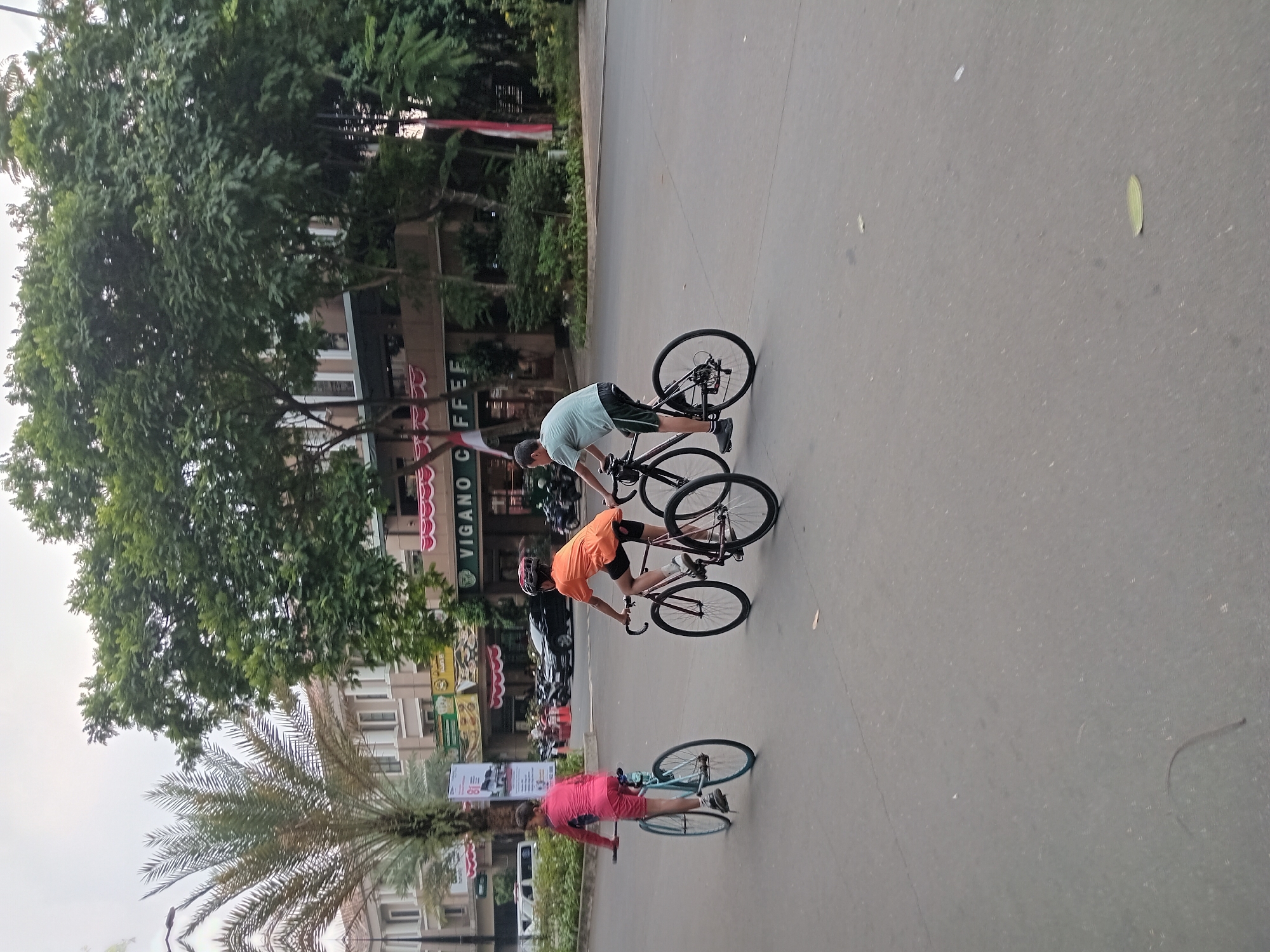 Three individuals are cycling on a paved road in Gunung Putri, Indonesia. The person on the left, appearing to be a child, is wearing a bright pink long-sleeved shirt and light blue shorts, riding a light blue bicycle. In the center, another child, wearing an orange t-shirt and black shorts with a red helmet, cycles a black bicycle with red accents. To the right, a person wearing a light blue t-shirt and black shorts is riding a black bicycle. In the background, there is a building with signs that read "VIGANO COFFEE" and other establishments. Several trees, including a palm tree, line the street. The sky is overcast. Vehicles, including cars and motorcycles, are parked in the background near the buildings. The road surface shows some wear and a few leaves are scattered on it.