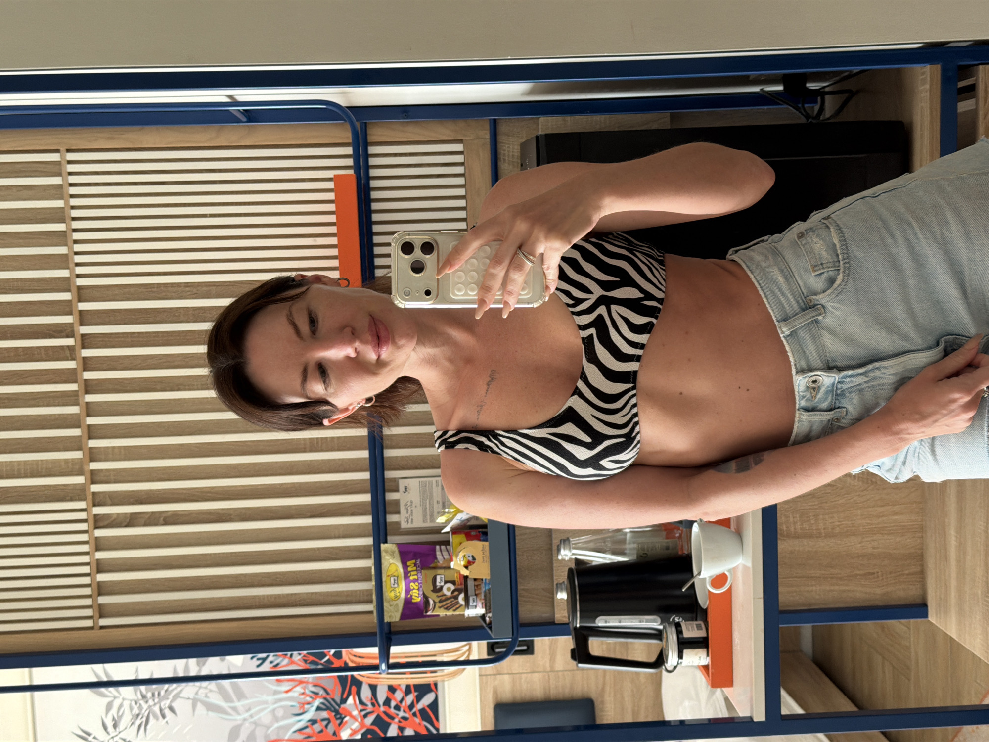 A woman is taking a selfie in a hotel room. She is wearing a black and white zebra print bikini top and light blue denim shorts. She has brown hair and is wearing hoop earrings and a ring on her left hand. She is holding a smartphone in her right hand. Behind her, there is a mirror reflecting a shelf with a black kettle, a bottle of water, a cup, a jar, and some packaged snacks. The room has wooden paneling on the wall. The background shows a glimpse of what appears to be a hotel room with furniture and decor. The image does not provide information about the city of Dong Ninh Hoa Ward, Vietnam.
