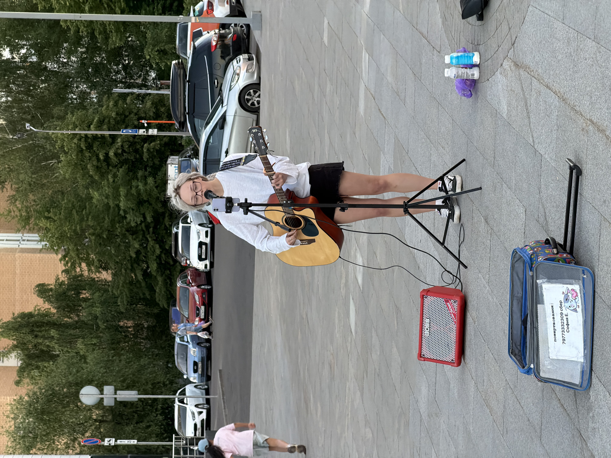 A young woman with blonde hair and glasses performs as a busker on a grey paved plaza in Moscow, Russia, playing an acoustic guitar and singing into a microphone mounted on a tripod with a smartphone attached. She wears a white long-sleeve shirt and black shorts. On the ground in front of her is a red BOSS CUBE Street amplifier. To her left, a blue soft-sided trolley bag is open, displaying a white sign with Cyrillic text that reads "пожертвования:" (donations:), a phone number "79773332309," "сбер" (Sber bank), and "София Е." (Sofia E.), accompanied by a cartoon image. To her right, a purple plastic bag and two white plastic bottles are visible. In the background, a young child in a pink shirt and light shorts runs to the left. Further back, two adults walk amidst a parking area filled with various cars, including a white Kia, a blue sedan, a red hatchback, a black SUV with a roof box, and a white Audi, some displaying Russian "777" region code license plates. Green trees, bushes, brick buildings, street signs, and lampposts frame the urban scene.
