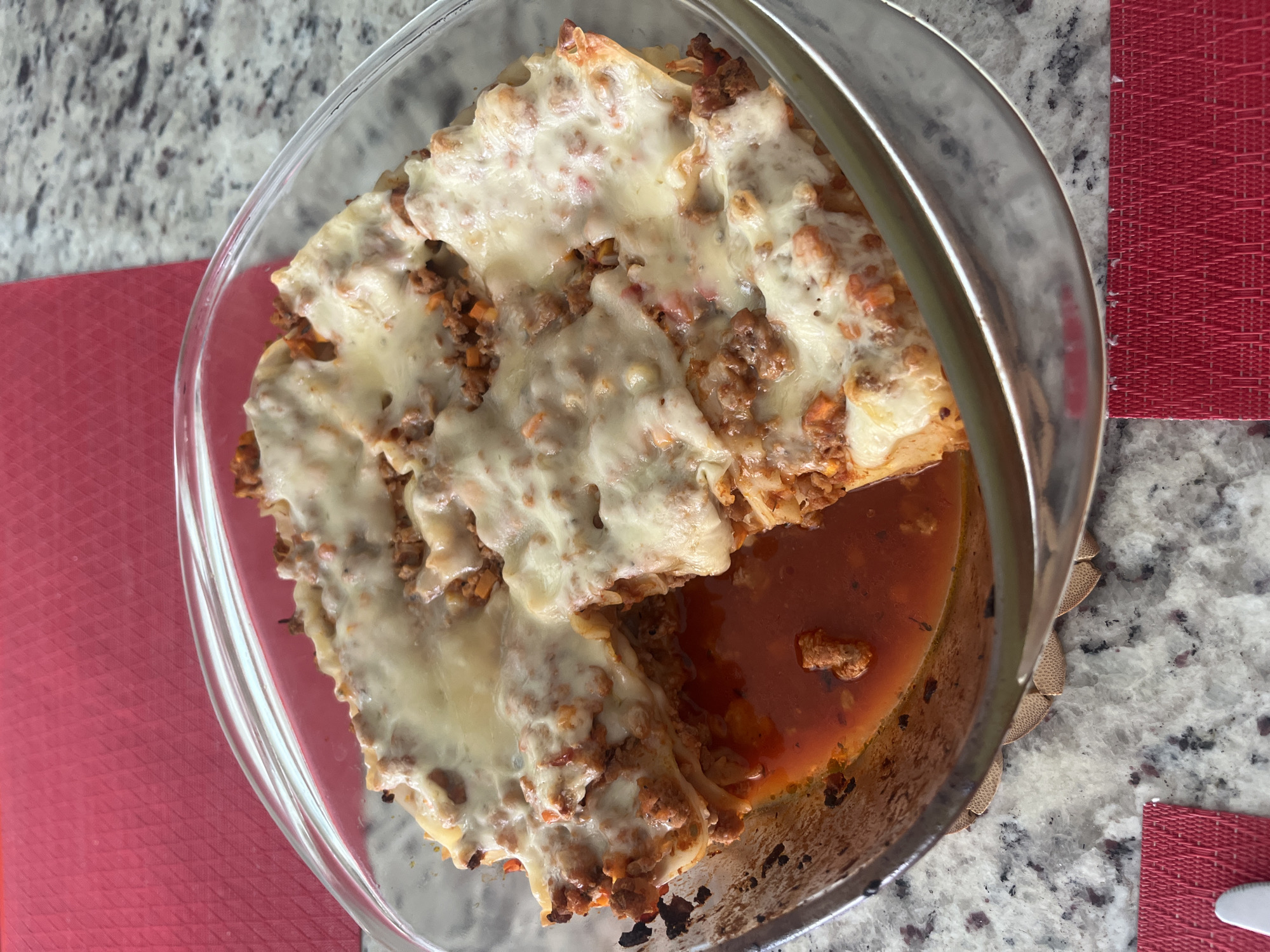 The image shows a glass baking dish containing lasagna on a countertop. The lasagna appears to have layers of pasta, meat sauce, and melted cheese. A pool of tomato-based sauce surrounds the lasagna in the dish. The countertop is a light gray speckled surface. Part of a red woven placemat is visible on either side of the dish. There is no evidence to suggest this image was taken in Barrio de Nuevo León, Mexico. No people, actions, or interactions are visible.