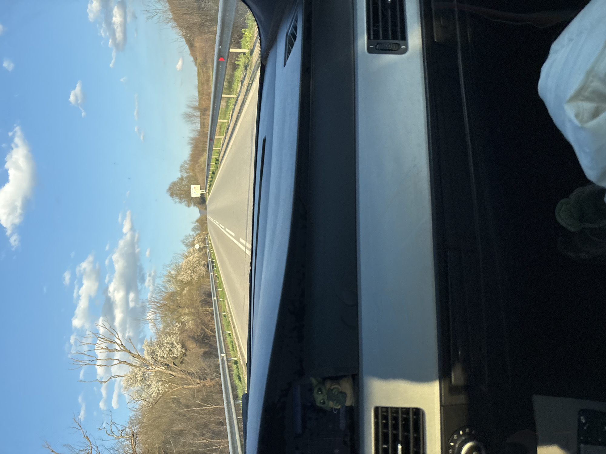 A view from inside a vehicle traveling on a road. The road is divided by a double white line. Beyond the road is a guardrail, trees, and a blue sky with white clouds. Inside the vehicle, the dashboard and air vents are visible. A figure resembling "Baby Yoda" is placed in a storage compartment. A white bag is partially visible in the foreground. The location is undetermined.
