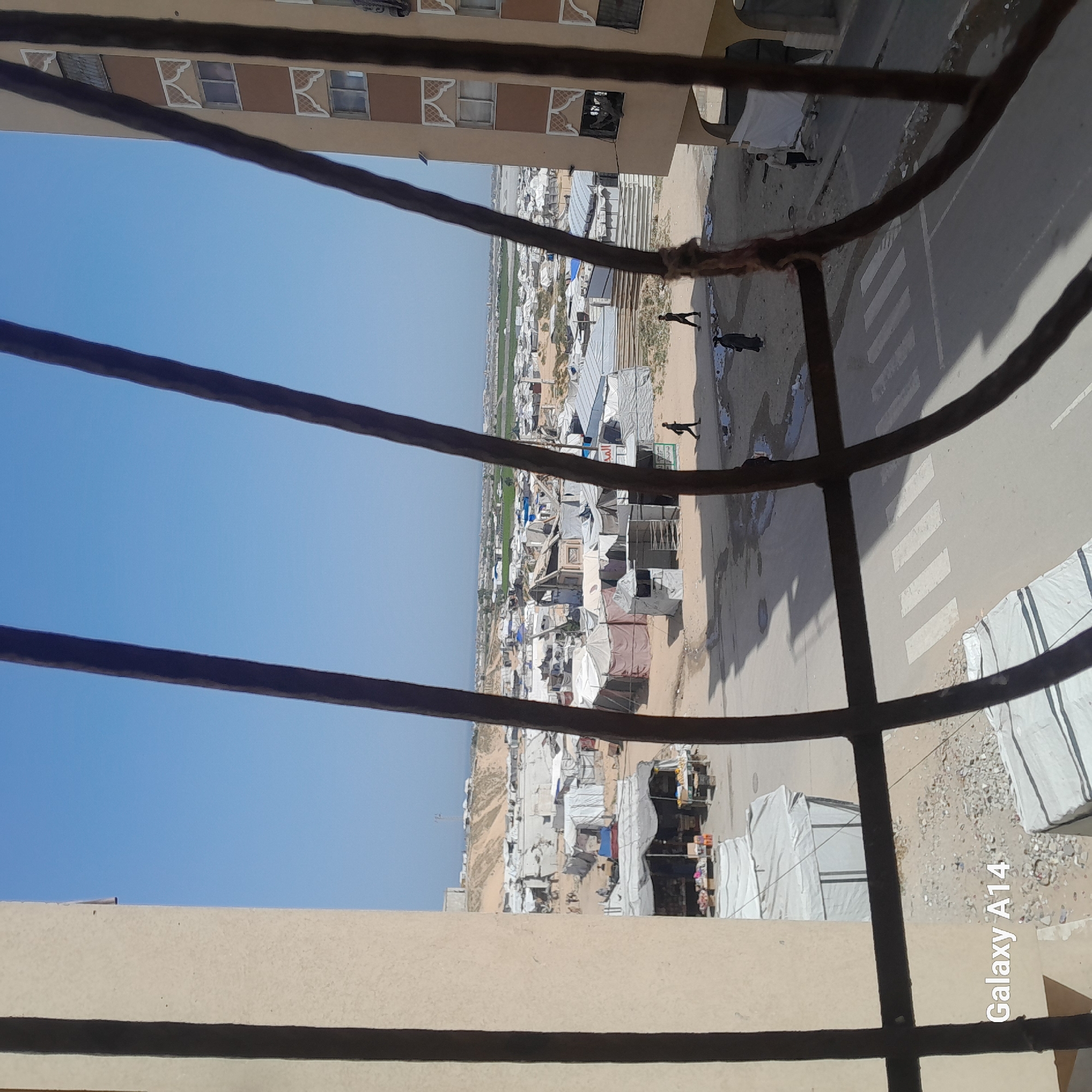 The view from a high vantage point through a wrought iron window looking down at a refugee camp next to a city, on a bright, sunny day. The camp is comprised of white tents and makeshift dwellings, with a few people walking along a road marked with a crosswalk. Across the road is a structure, possibly a building, with a sign in Arabic. Farther out is a green field bordering the urban landscape.