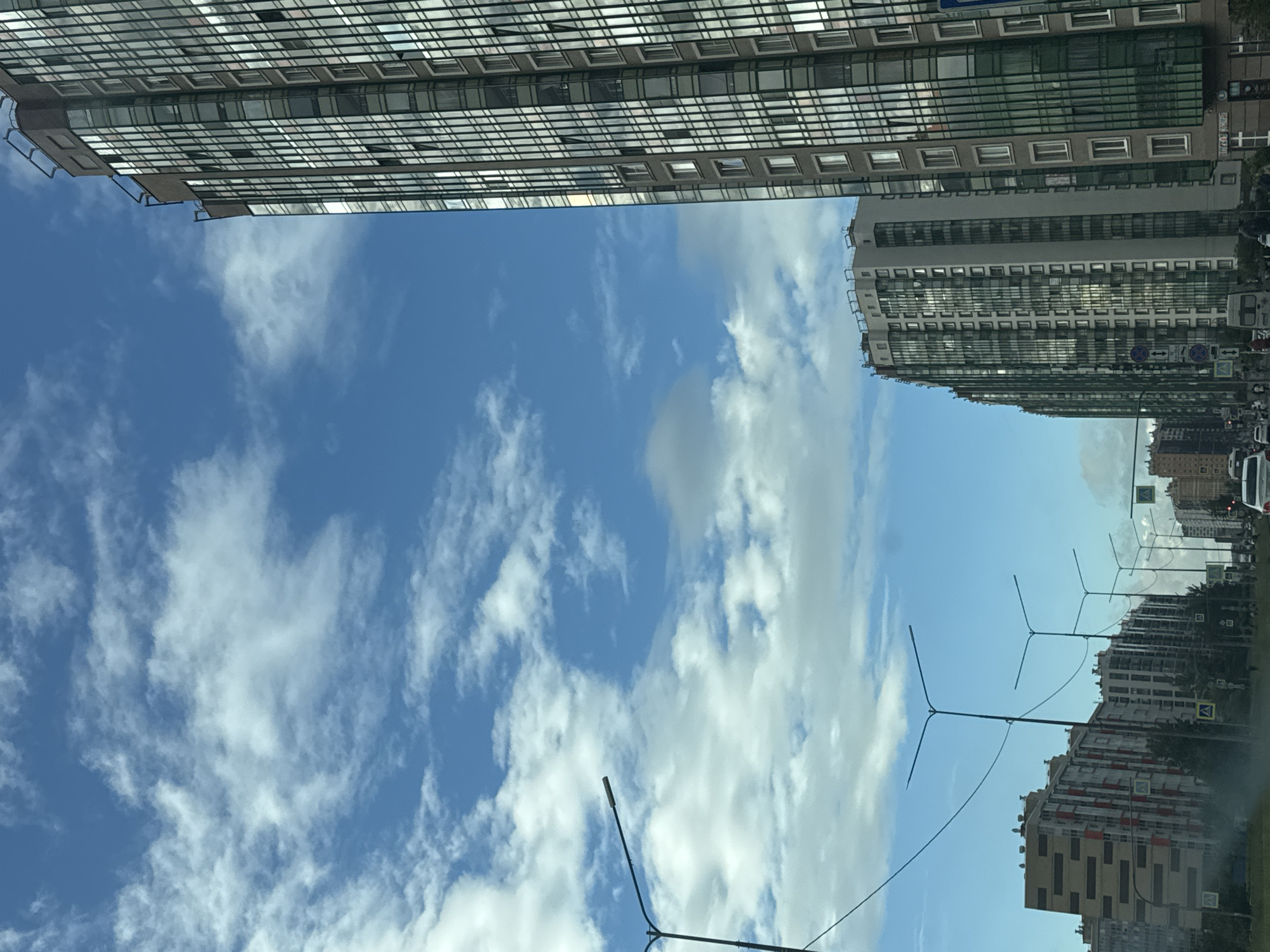 This media file shows a view of apartment buildings against a blue sky with scattered clouds. The image is oriented vertically, as if taken from the perspective of someone looking up.
The most prominent subjects are the tall apartment buildings, characterized by their repetitive window patterns. One building on the left appears to be a modern high-rise with a facade of glass and reflective surfaces, while other buildings on the right show more variation in color and design, with some featuring balconies. Streetlights with a distinctive multi-branched design are visible between the buildings.
The scene depicts an urban environment. There are no people clearly visible. The activity, if any, is not evident, and the image appears to be a static shot. The time of day seems to be daytime, given the bright sky and natural light. The weather appears to be partly cloudy, with a mix of blue sky and white clouds.
Notable details include the architecture of the buildings and the design of the streetlights. There are no clear location cues to pinpoint a specific place, other than the provided context of "Unknown, Russia." No emotions are conveyed. There is some very small, illegible text on signs in the distance, but it is not discernible.
