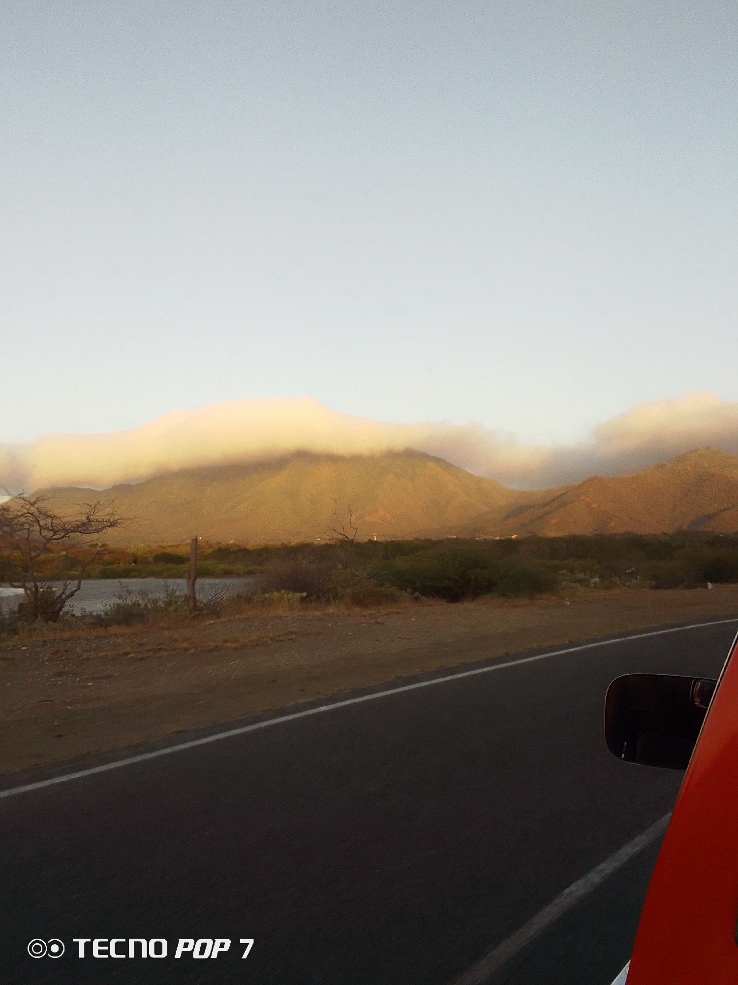 The photograph shows a mountainous landscape under a hazy sky, likely taken from a moving vehicle. A road with a white dividing line occupies the lower portion of the frame, with a portion of an orange car visible on the right. The background features a range of mountains, partially obscured by a low-lying cloud formation that appears to envelop their peaks. Vegetation, including shrubs and trees, is visible in the foreground, suggesting a rural setting. The camera used to take this photograph is a TECNO POP 7. The location is likely near Altagracia, Venezuela.