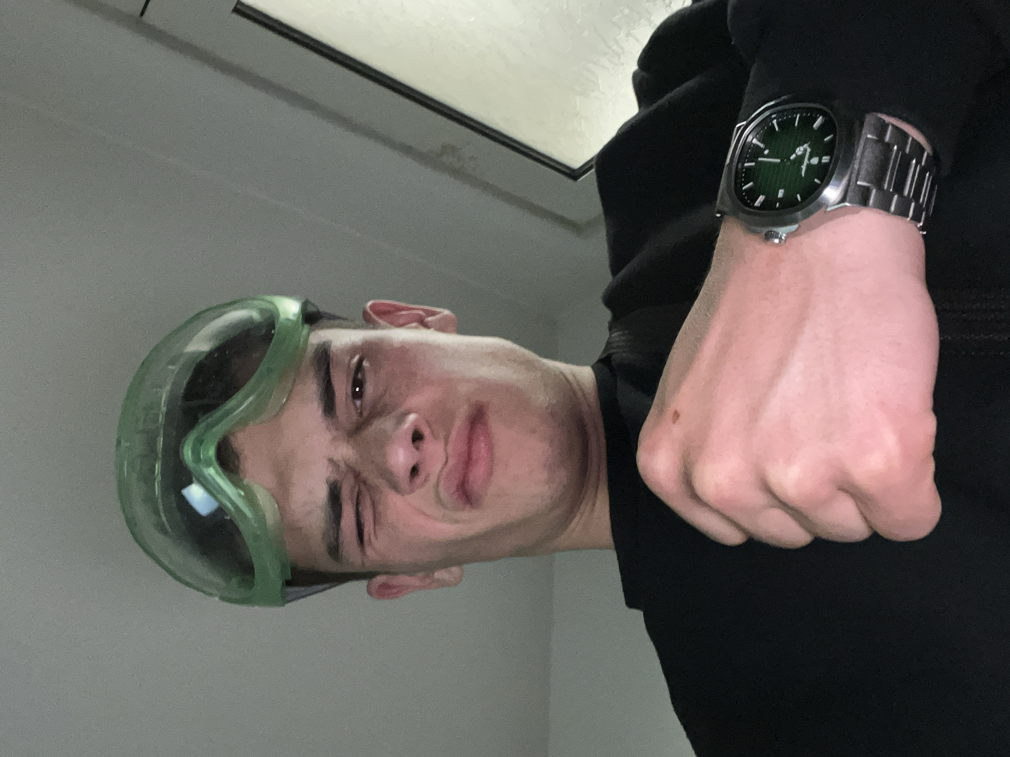 A person wearing green goggles and a silver watch with a green face is posing for a selfie in an indoor location, with a window visible in the background. The person is wearing a black garment. The location appears to be indoors. There is no information to confirm the location is in Karavelovo, Bulgaria.