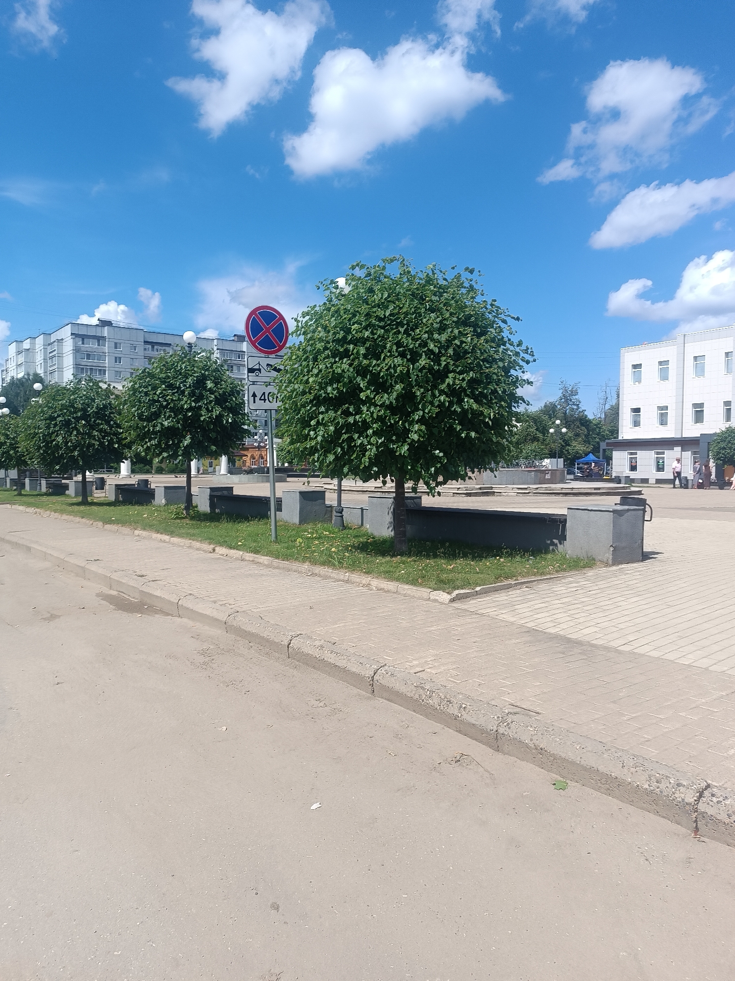 The image captures a street scene in Chaikovsky, Russia, on a sunny day with a bright blue sky filled with fluffy white clouds. The foreground features a paved road transitioning into a brick-paved sidewalk bordered by a concrete curb. To the left of the sidewalk, a strip of grass separates it from a row of well-maintained, spherical trees. Behind the trees, there are concrete benches or planters.
A traffic sign is prominently displayed on a pole. The sign has a red circle with a blue background and a red diagonal line, indicating "No Stopping." Below this, another sign shows a tow truck icon and a number "40" with an arrow pointing upwards, suggesting that stopping is prohibited and vehicles may be towed if they stop for longer than 40 meters.
Further back, on the left side of the image, a multi-story apartment building with a gray facade and white window frames is visible. On the right side, a modern white building with multiple windows stands, possibly a public building or office. In the middle ground, there are more trees, some greenery, and what appears to be a small structure covered by a blue tent. A few people are visible walking in the distance near the white building. The overall impression is a clean and organized urban environment.