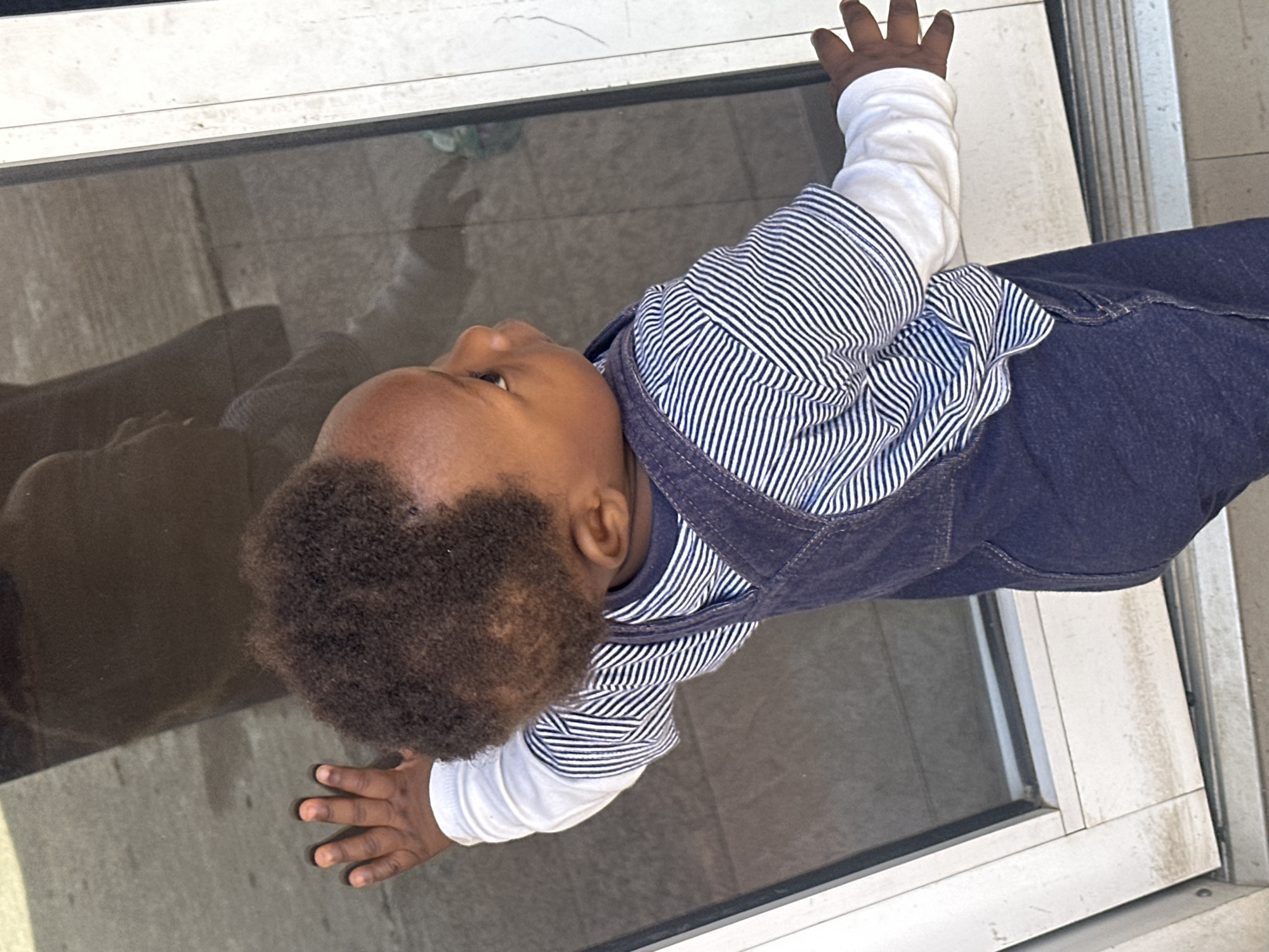 An infant is standing and looking out a glass door, their right hand resting on the glass. The infant is wearing a white long-sleeved shirt under a navy blue striped shirt and navy blue overalls. The infant's reflection is visible in the glass, which appears to be a sliding glass door to an outdoor area. The ground outside the door is concrete. The scene is outdoors, likely in a residential or public area in the city of Gura Top, Nigeria.