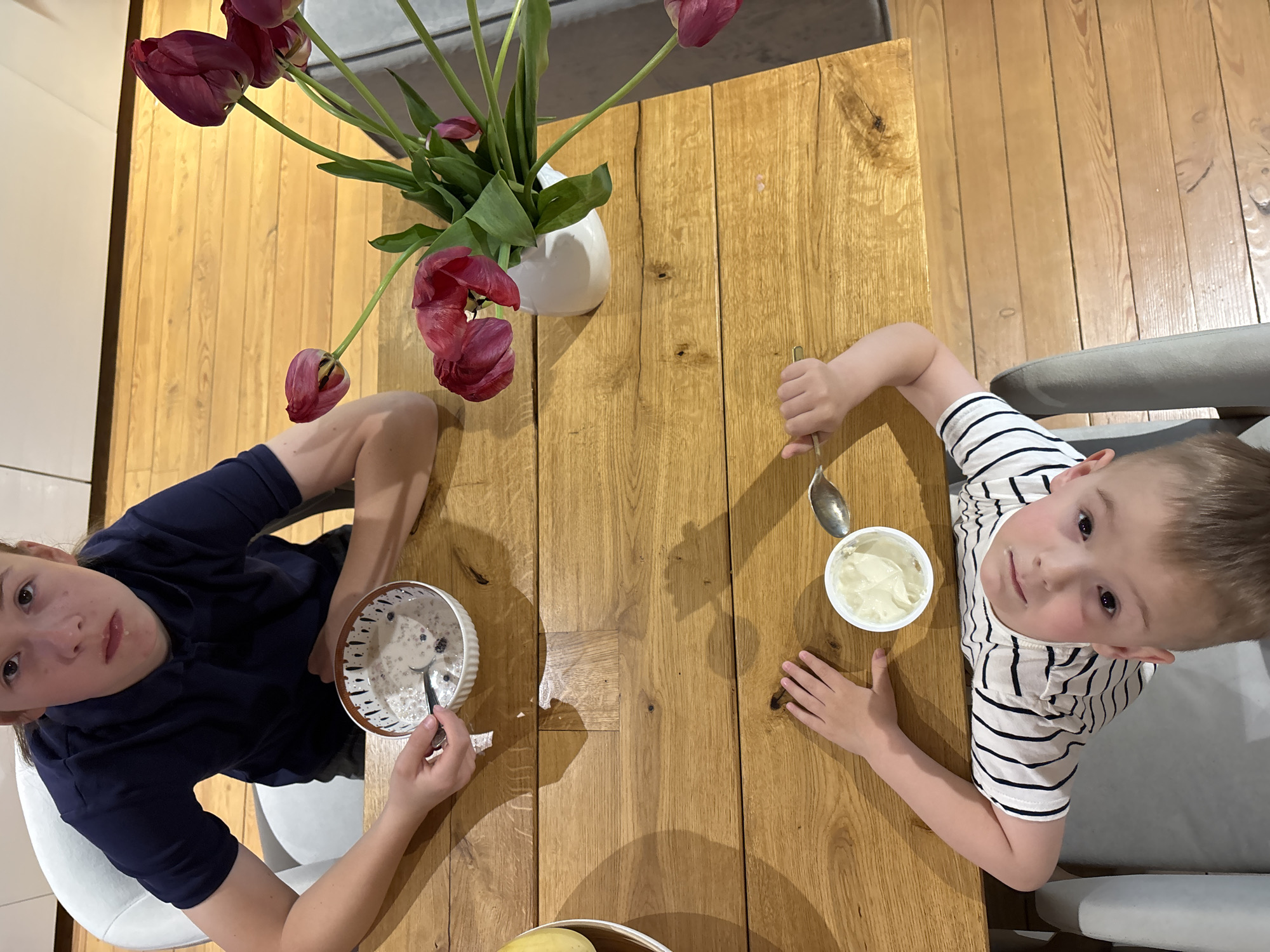 Two children are seated at a wooden table eating from bowls. A vase of red tulips sits in the center of the table. One child is wearing a navy blue shirt and is eating from a bowl with dark markings around the rim. The other child wears a striped white and navy shirt and has a white bowl. The location cannot be determined to be Chaikovsky, Russia from the image.