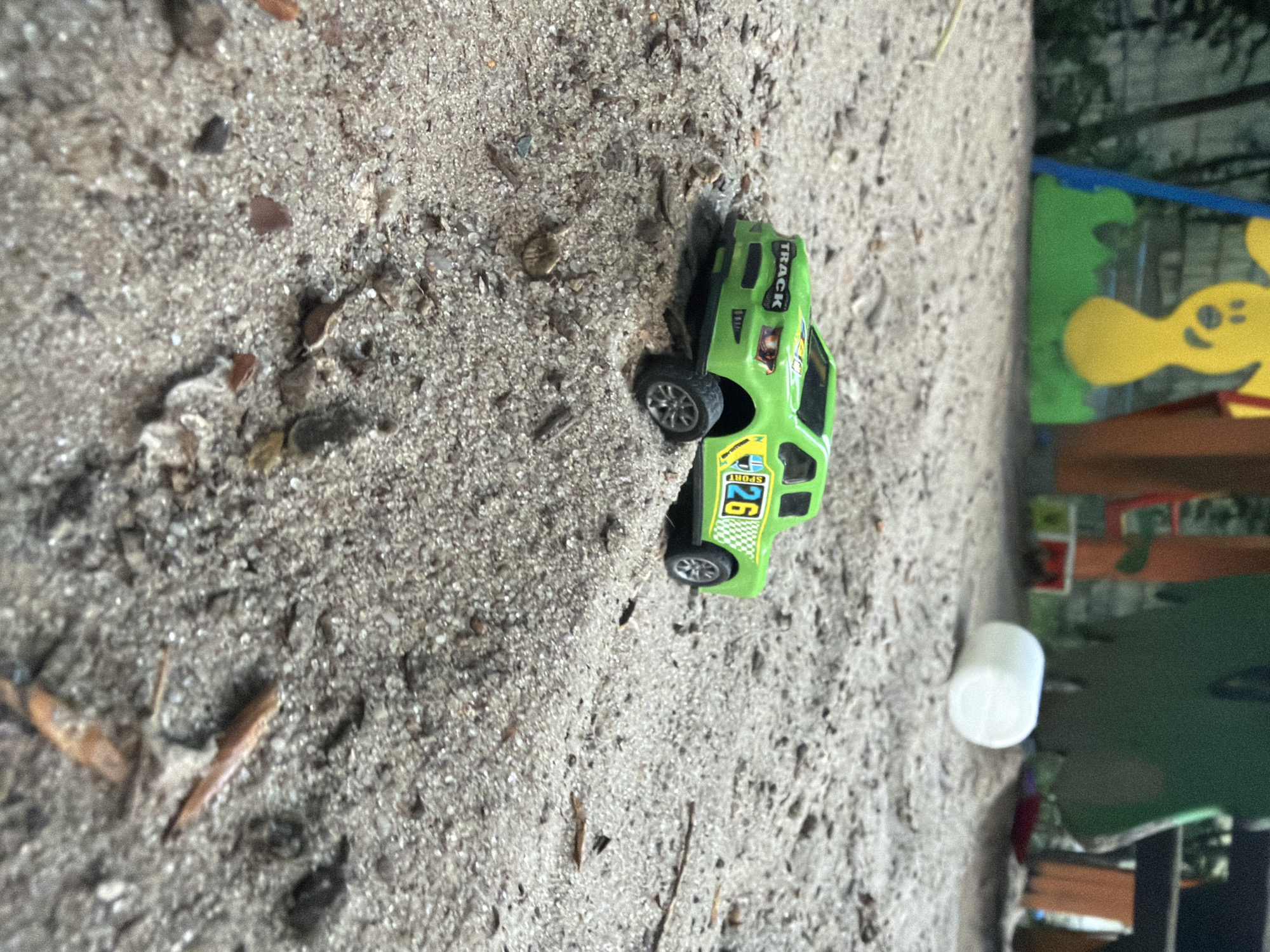 In Kyiv, Ukraine, a miniature green off-road toy truck with the number 26 on its side is positioned on a sandy surface. The truck features the word "TRACK" above the front grille. The environment appears to be an outdoor play area, with blurry, colorful structures in the background and scattered debris on the sand. A white cylindrical object is also visible in the sand to the right of the truck.