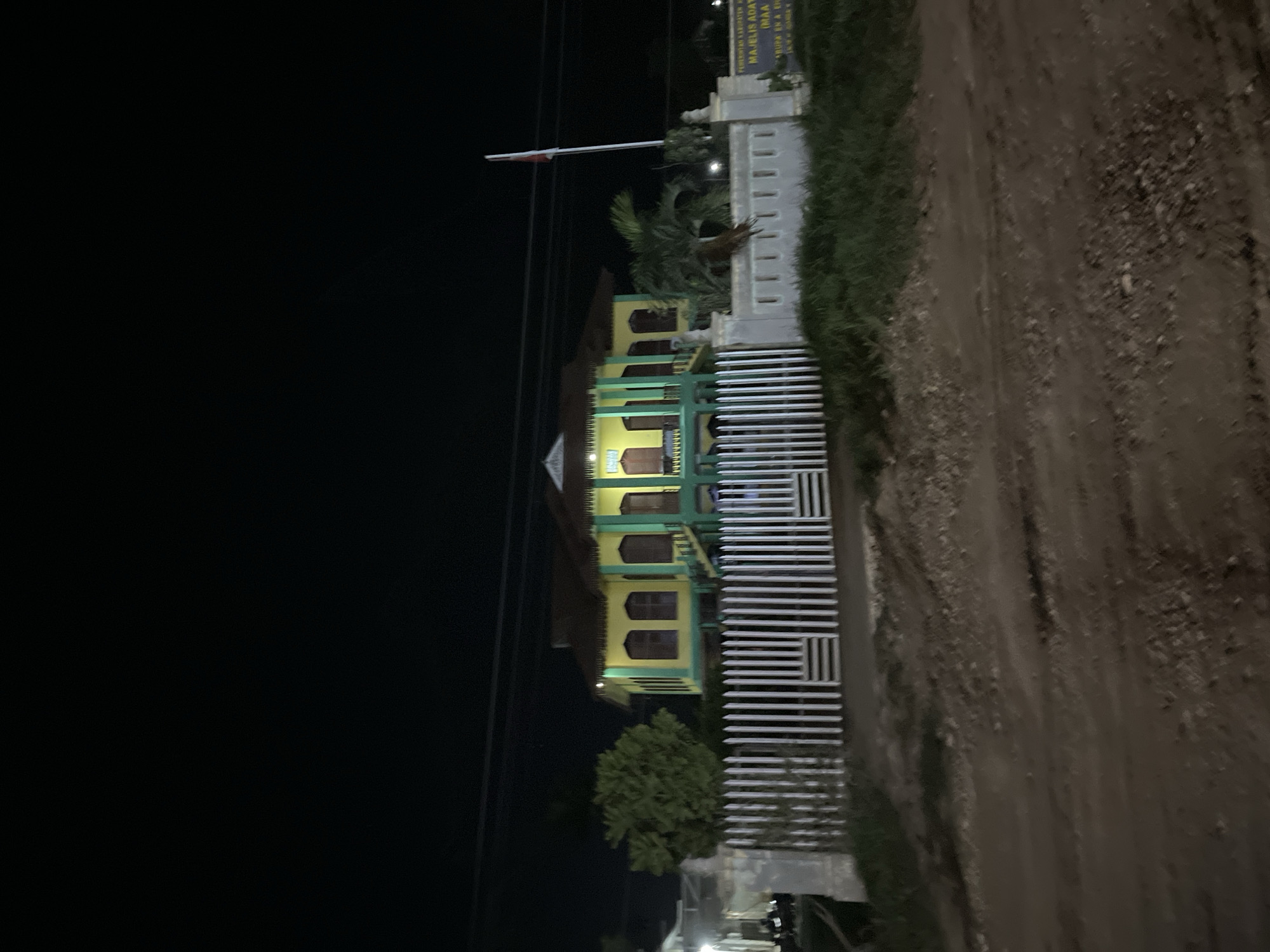 The image depicts a multi-story building, likely a school or government institution, at night in Bundar, Indonesia. The building is primarily yellow with green trim and features multiple windows and balconies. A white fence with horizontal bars separates the building from a dirt path on the right side of the image. A flagpole with a red and white flag stands to the left of the building. The scene is illuminated by artificial lights, suggesting it is evening or night. A sign is visible above the fence, partially obscured by darkness and greenery, with text that appears to be in Indonesian. The environment is dark, with a clear sky, indicating no precipitation. The overall mood is quiet and still, characteristic of a nighttime scene in a semi-urban or rural Indonesian setting.
