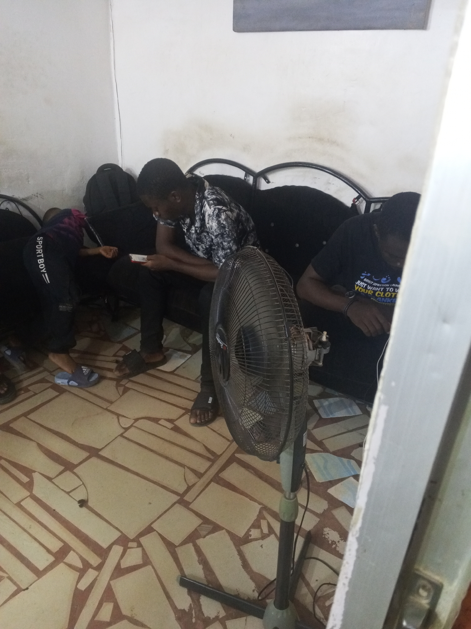 A group of three people are sitting on a black couch indoors in what appears to be a room in Gwong, Nigeria. One person is dressed in black pants and a patterned black and white shirt and is looking down at an object in their hands. Another person is lying down on the couch, wearing black pants with the word "SPORTBOY" printed on the side, and blue sandals. The third person is wearing a black t-shirt with printed text. In the foreground is a standing fan. The floor has a pattern of geometric shapes. A black backpack is placed on the couch in the background.