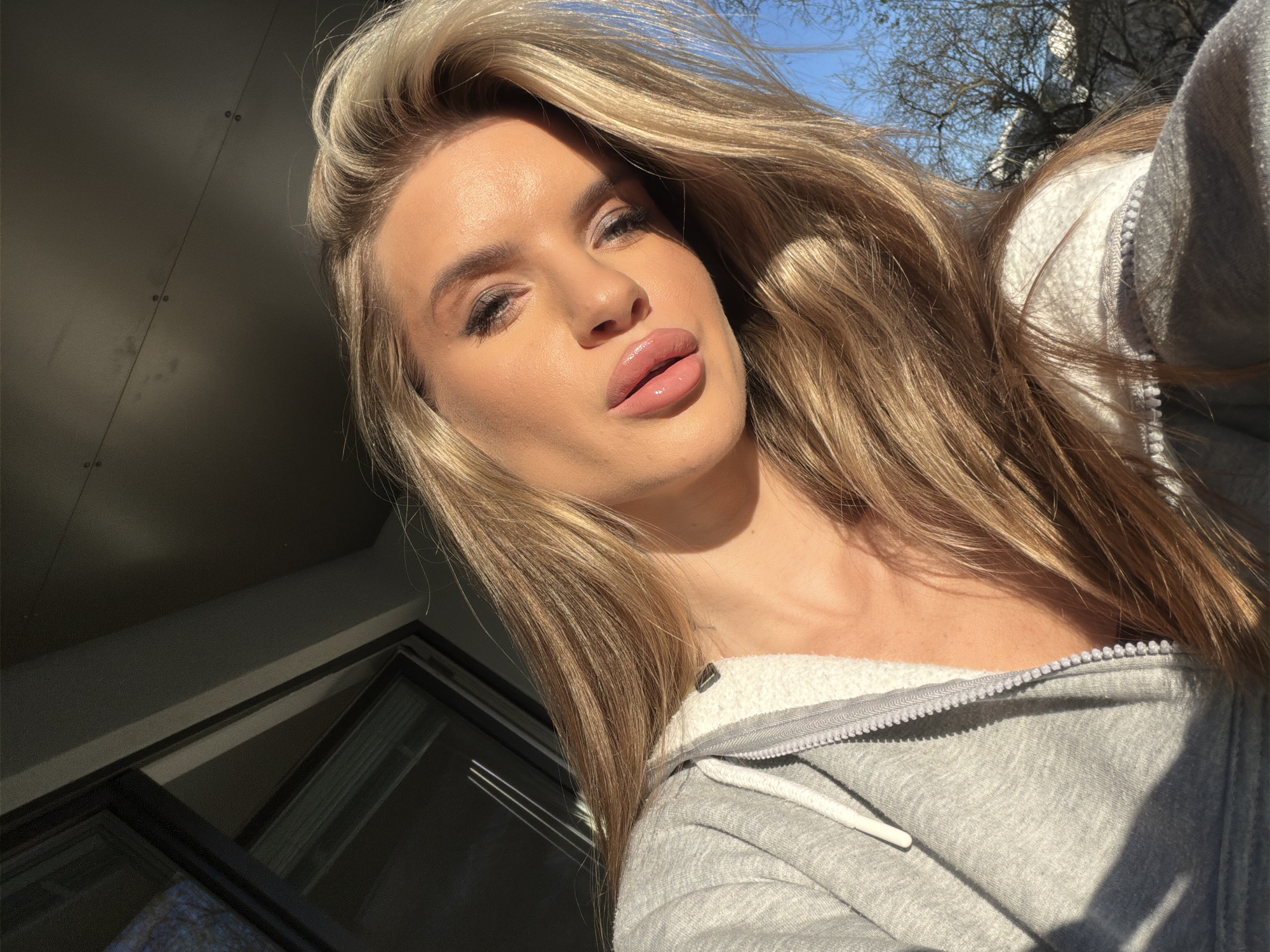 A medium shot captures a blonde woman with fair skin and full lips, wearing a grey zip-up hoodie, positioned in front of a dark, modern building facade, possibly a residential apartment building. The sunlight bathes her face and hair, creating strong highlights and shadows. In the background, the branches of a tree are visible against a clear blue sky. The woman appears to be taking a selfie. Given the architectural style and prevalent use of modern building materials, the location might be inferred to be in a contemporary residential district of Berlin, Germany.
