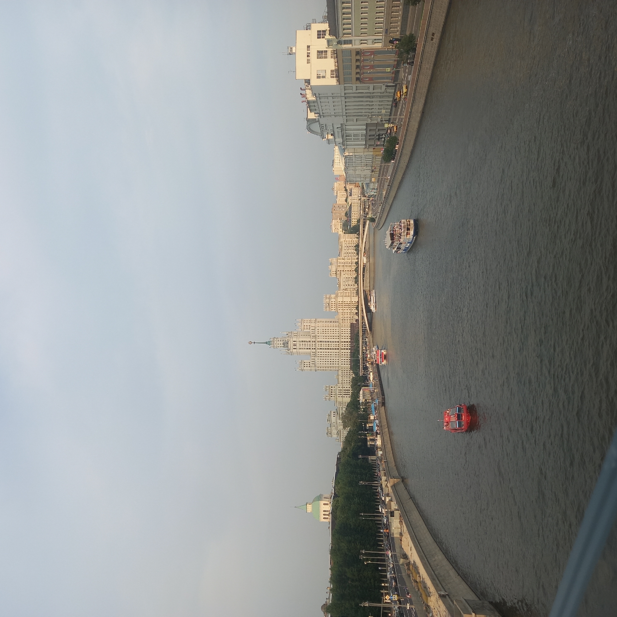 The image depicts a wide, calm river flowing through a city. Several boats are visible on the water, including two red passenger boats and two larger white cruise boats. The riverbanks are lined with concrete embankments and buildings. On the left bank, a road with traffic and trees runs alongside the embankment. In the distance, a prominent white skyscraper with a spire dominates the skyline, flanked by other buildings. On the right bank, a row of light-colored buildings with a modern architectural style runs along the river. The sky is overcast with a light haze. The overall scene is a cityscape with a focus on the river and the architecture along its banks.