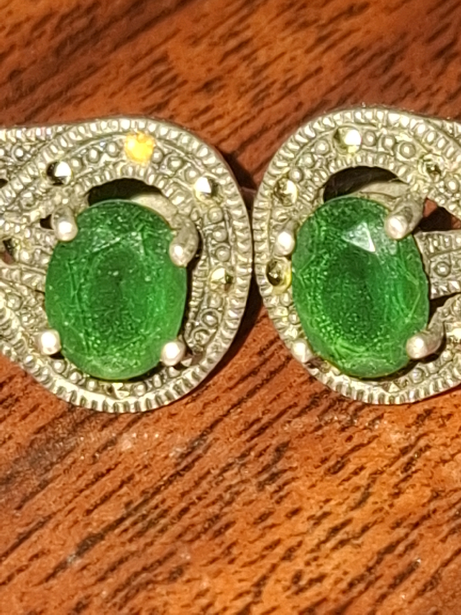 The image shows a close-up of two ornate silver earrings, each featuring a large, oval-shaped green gemstone. The earrings are detailed with intricate filigree work and small, round, white stones that appear to be marcasite, as well as tiny yellow accents. The earrings are resting on a textured, reddish-brown surface, likely wood, with visible grain. No people or actions are depicted in the image. The image does not provide any information about the city of Almaty, Kazakhstan, or any specific location within it.