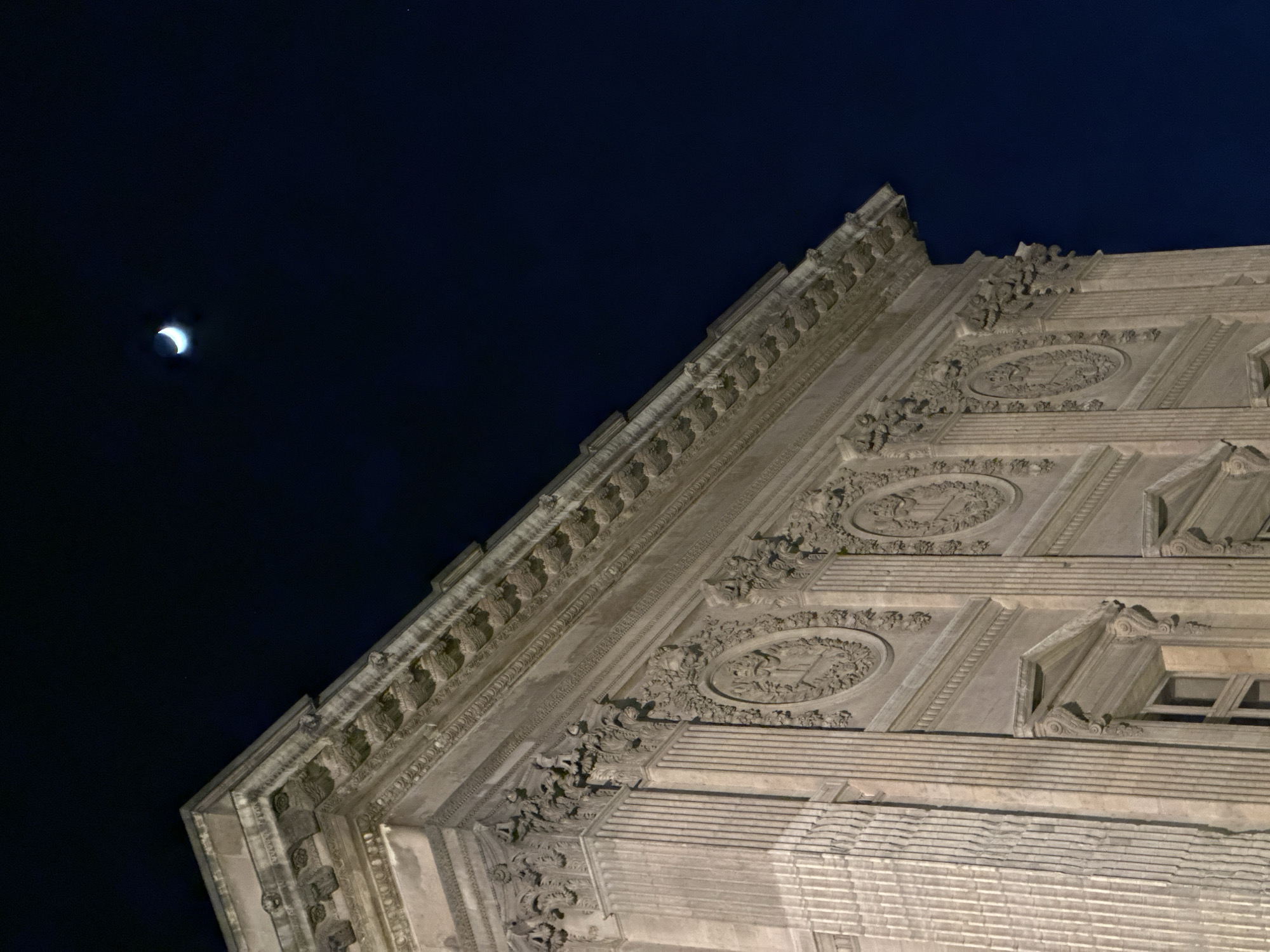 The image captures a portion of an ornately decorated building facade against a night sky with a crescent moon. The building features elaborate architectural details including moldings, friezes, and decorative panels. The facade appears to be constructed of stone or a similar material. The sky is dark, with the moon visible in the upper left portion of the frame. The location is unidentifiable from the image, though the architectural style suggests a European city. The building's design and the presence of the moon are the only elements present; no people, animals, or other specific objects or actions are visible. The environment is a nighttime urban setting.