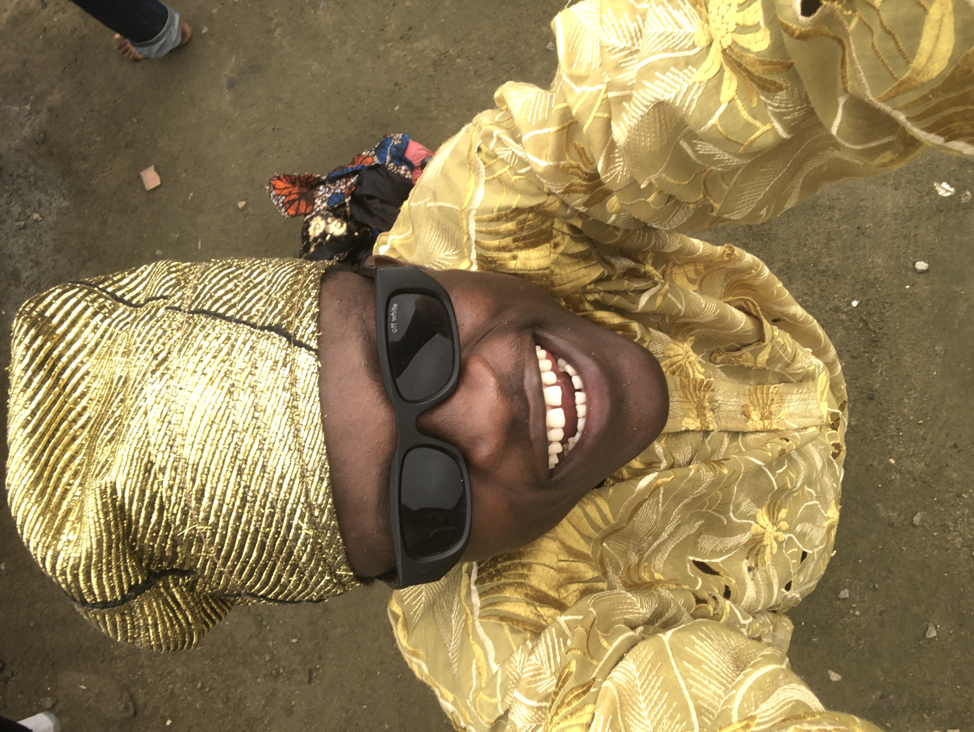 A close-up, high-angle shot shows a person wearing a gold patterned headwrap and a gold patterned garment, smiling at the camera. The person is also wearing black sunglasses with the text "off white" visible on the left lens. In the background, a pile of colorful fabric is visible, and to the right, a person's legs are visible wearing blue jeans and bare feet. The ground appears to be dirt or gravel. The location is described as Ado, Nigeria.