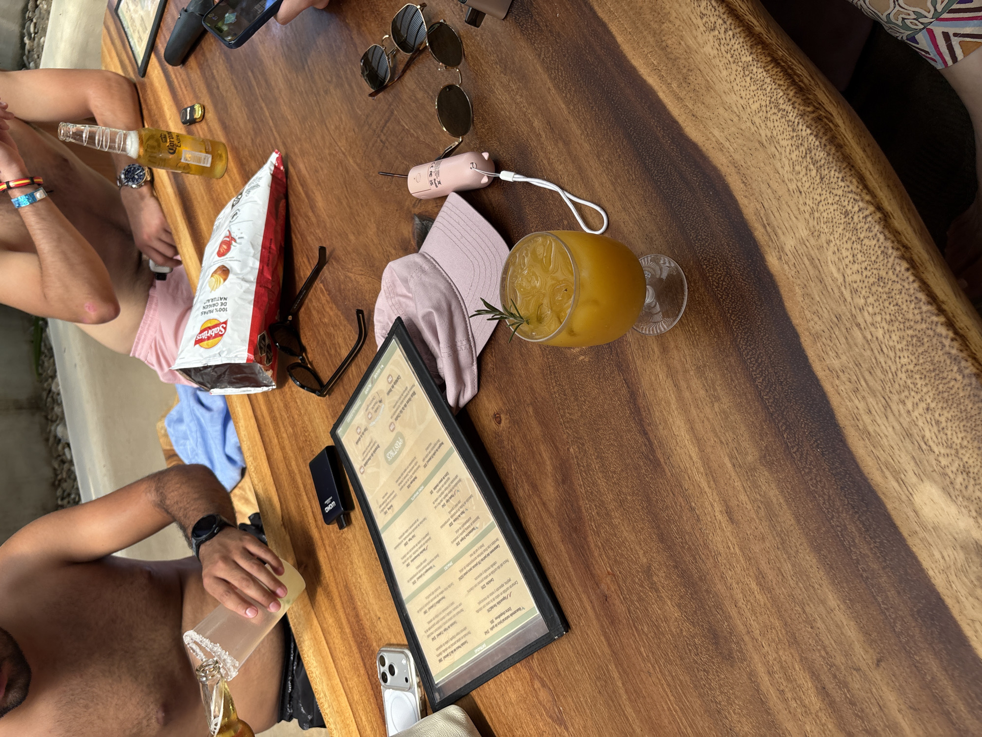 A wooden table is shown from above, populated with various items suggesting a leisure setting. A glass of amber-colored liquid with ice and a sprig of rosemary sits to the right. Next to it is a pink baseball cap and a small, pink electronic device. Further left, there is a pair of sunglasses, a bag of Sabritas potato chips, and another pair of sunglasses. A menu is propped up on the table. A man is pouring a beer into a glass. The presence of a phone, a power bank, and a wrist watch suggests the image was taken in Mexico, city of Ministerios de Amor Eterno Asociación Cultural.