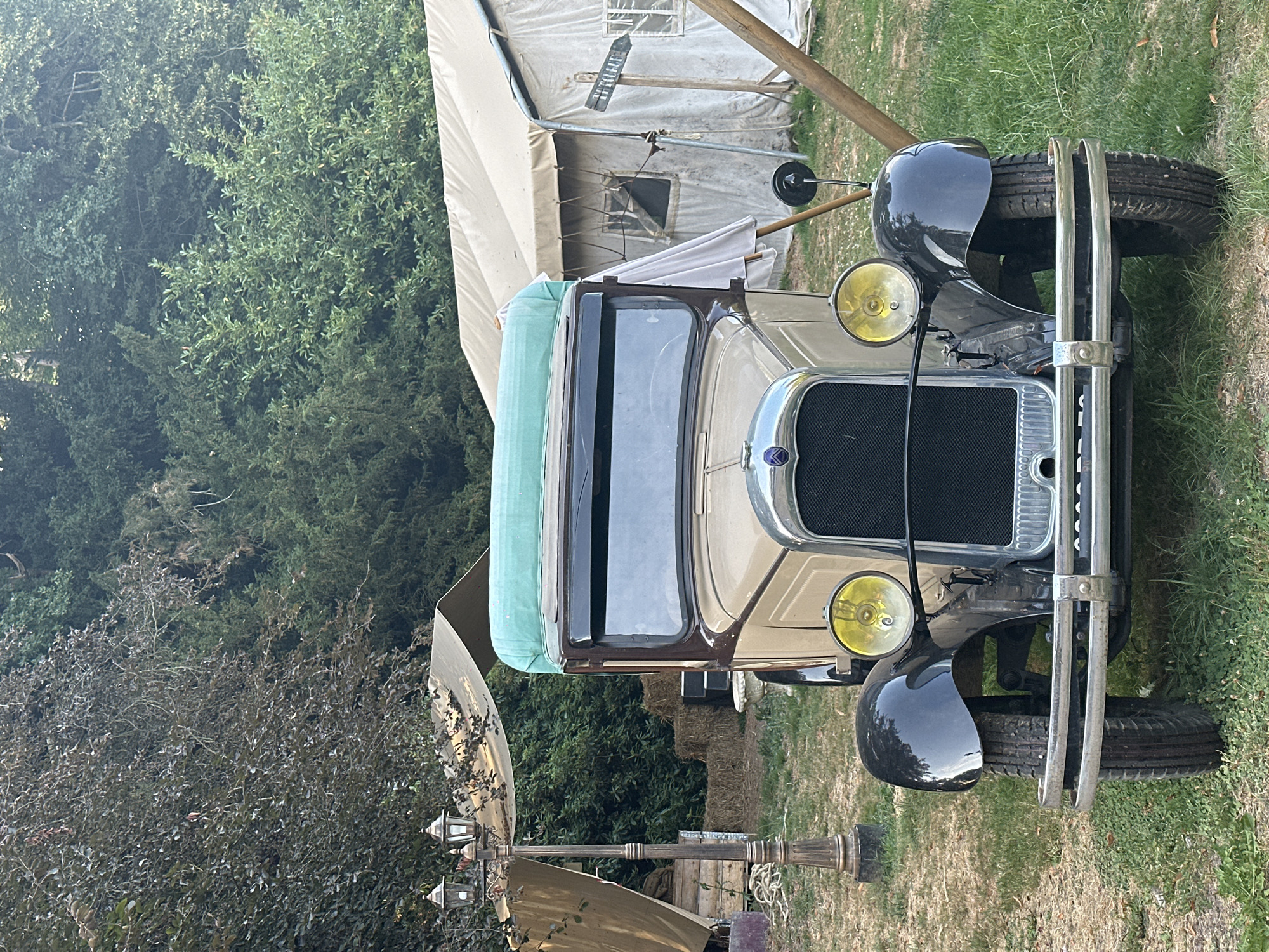A vintage, two-tone beige and brown truck with a green tarp draped over its roof is parked on a grassy area. The truck has a chrome grille, two yellow headlights, and a chrome bumper. It is facing forward, with its front wheels on the grass. In the background, there are several canvas tents, hay bales, and trees. A vintage lamppost with three lights stands to the left of the truck. A wooden sign with arrow pointing right, with the word "TUTTI LE", is attached to a wooden pole to the right of the truck. The scene appears to be set up for an outdoor event or festival. The location is Saint-Vaast-du-Val, France. There are no people visible in the image.