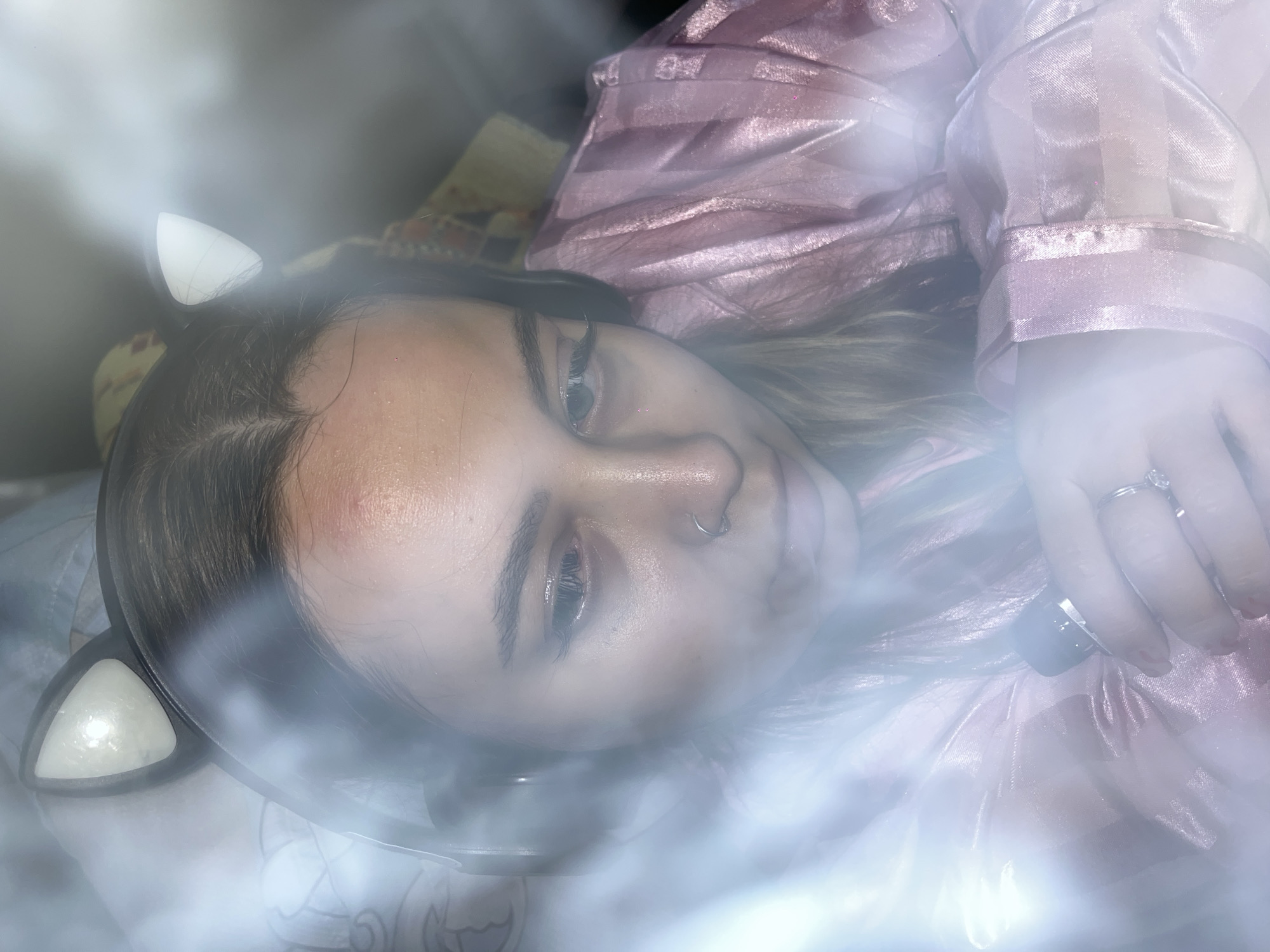A female with a nose piercing and a ring on her finger is lying on a bed in Novopokrovskaya, Russia. She is wearing cat ears and a pink striped pajama top. She is holding an object in her hand, obscured by light. The scene is filled with fog or smoke.