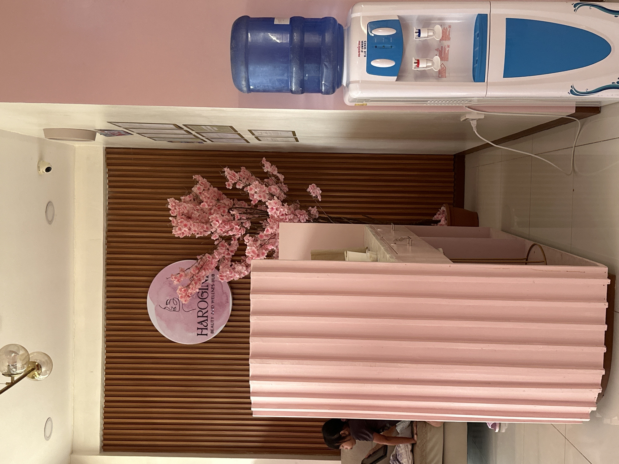 This image depicts the interior reception area of the "HAROGINA BEAUTY AND WELLNESS HUB" located in Samal, Philippines. The scene is dominated by a pink and wood aesthetic.
In the foreground, a tall, fluted, light pink reception counter stands prominently. Behind it, a child with dark hair, wearing a dark shirt, is seated on a light-colored couch, facing left and partially obscured by the counter. A purple shoe is visible near the child's feet on the tiled floor. To the left, a light-colored countertop extends with what appears to be a sink.
The background features a wall composed of vertical wooden slats. Centered on this wooden wall is a circular pink sign with a minimalist line drawing of a face in profile. The text "HAROGINA BEAUTY AND WELLNESS HUB" is printed below the drawing in a dark font. To the right of the sign, a large artificial tree adorned with numerous pink cherry blossoms stands, extending upwards and outwards. Its brown pot is visible at the base.
To the right of the main reception area, a plain pink wall extends from floor to ceiling. Adjacent to this, on a cream-colored wall, a security camera (CCTV) is mounted on the ceiling. Below it, a partially visible cream-colored round wall clock is mounted, followed by a vertical arrangement of several framed documents. A white and blue water dispenser, branded "AMERICAN POWER BLUE SERIES," is positioned against the pink wall. It features a large blue water bottle on top, two spigots (red and blue), and a blue collection tray. A white power cord from the dispenser extends along the light-colored tiled floor to a wall outlet on the cream-colored wall.
The ceiling is white with recessed lighting. A modern light fixture featuring three spherical glass globes suspended from a brass-toned rod is visible in the upper left of the frame.