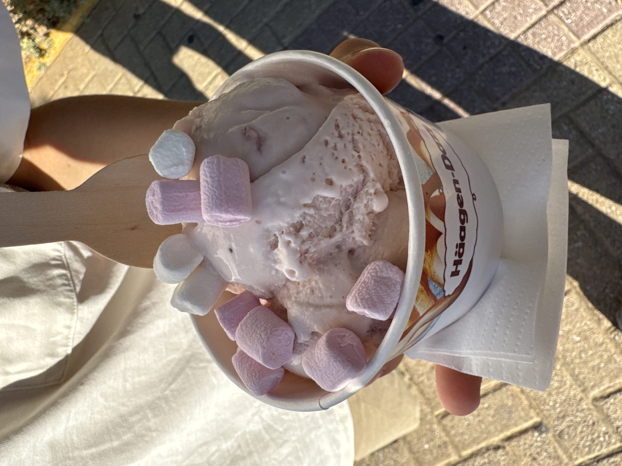 An overhead close-up shot shows a cup of strawberry ice cream topped with marshmallows. The ice cream is a pale pink color and is melting slightly. There are several small, puffy marshmallows, some pink and some white, scattered on top of the ice cream. The cup has the Häagen-Dazs logo visible. A wooden spoon is partially visible at the top of the frame. The cup is being held by a person, with their hand visible on the right side. The background shows a sunny outdoor scene with cobblestone paving and shadows cast by sunlight. The image does not provide any specific information that would indicate the location is Moschato, Greece.
