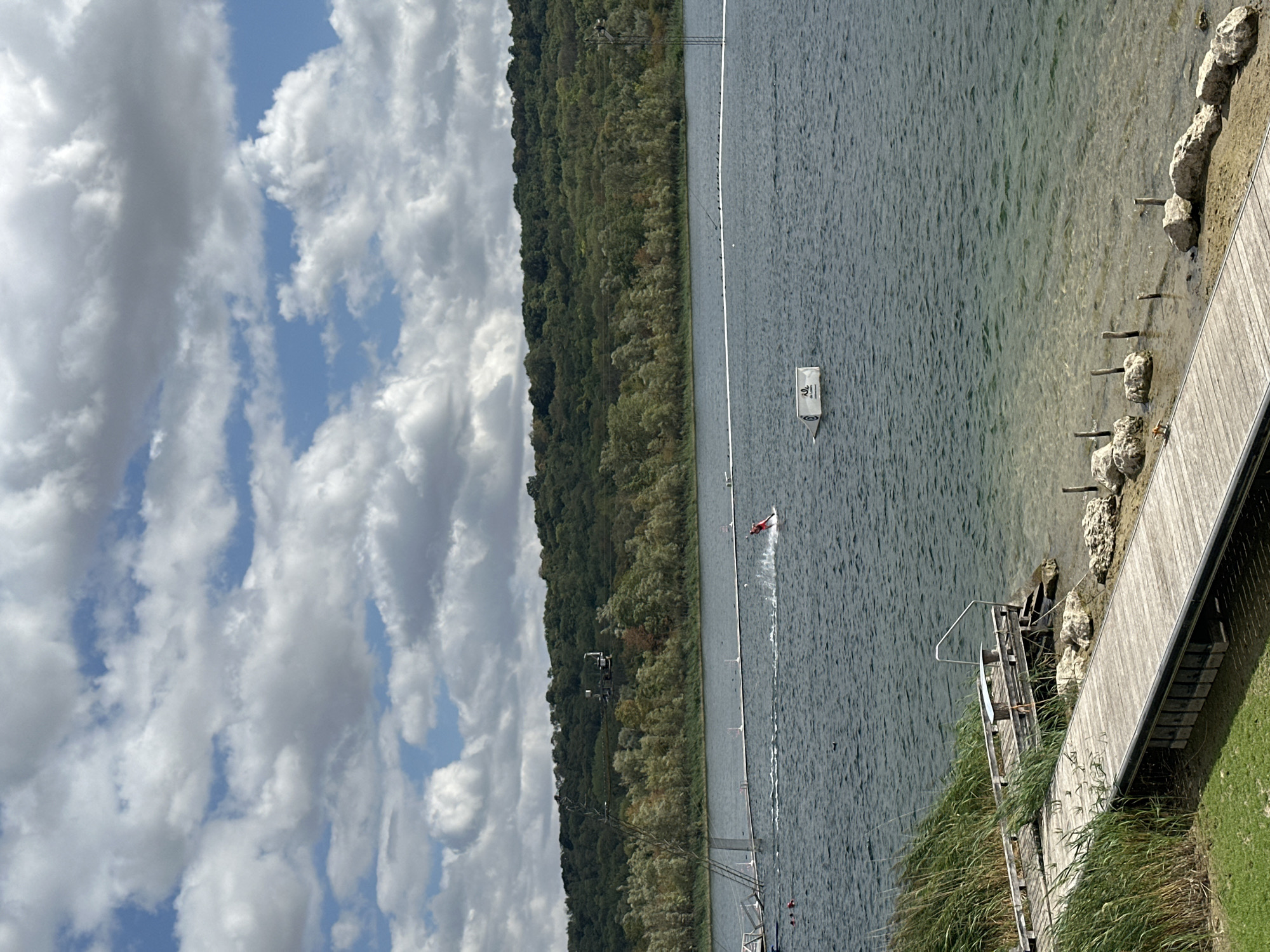 The image depicts a daytime scene at the watersports complex located in Jablines, France. The sky is filled with a mix of white and grey clouds against a blue sky. In the foreground, a wooden boardwalk extends into the water. To the left of the boardwalk, tall green reeds are visible. The water is choppy, with a visible wake. In the mid-ground, a person in a red suit is waterskiing, leaving a white spray behind them. Further out on the water, a white rectangular float is visible. In the background, a densely wooded hill forms the landscape. The overall impression is one of an active recreational area on a partly cloudy day.