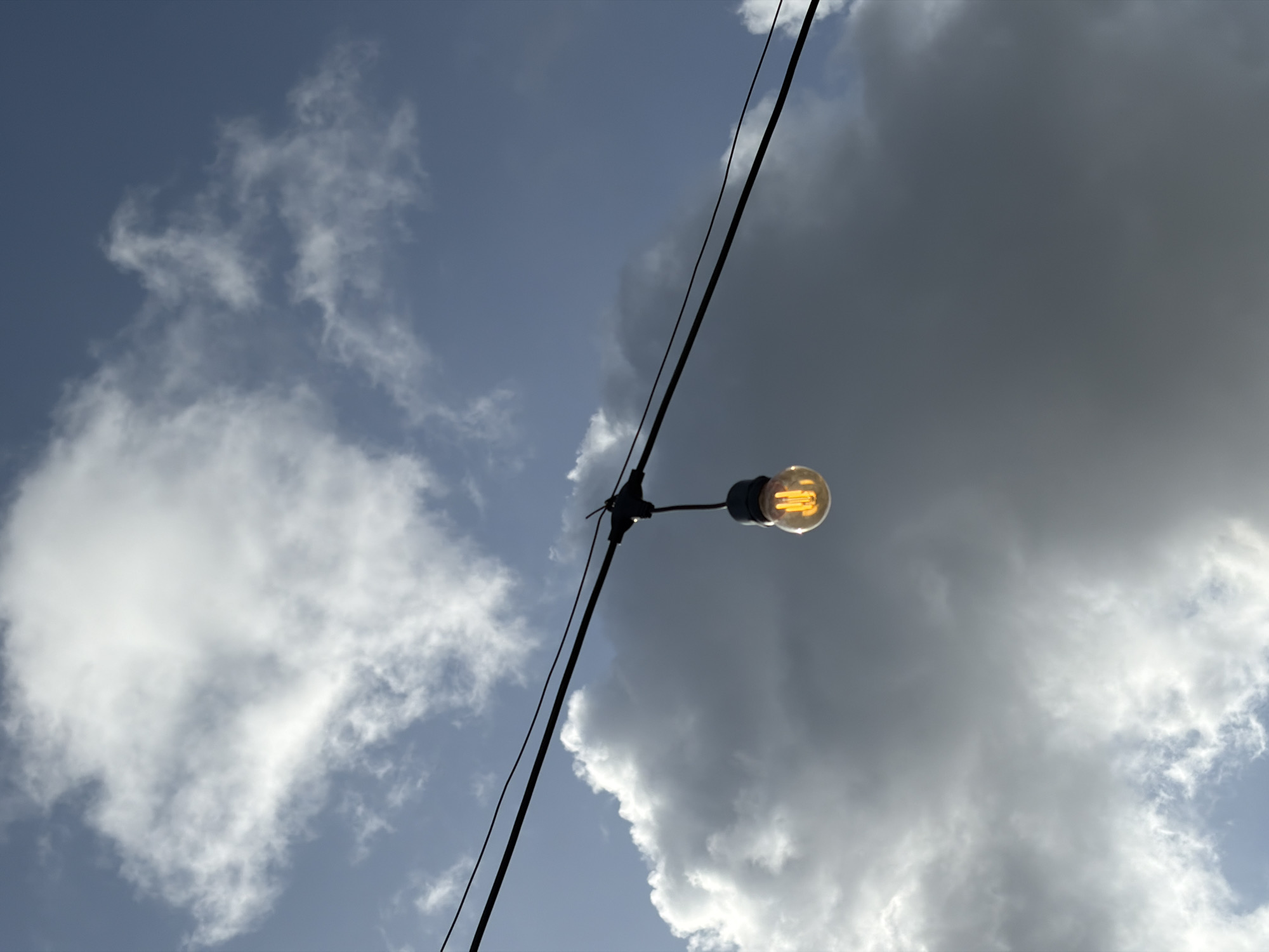 A low-angle shot captures a lightbulb suspended from a wire against a backdrop of a partly cloudy sky. The lightbulb is illuminated, displaying a warm yellow glow. The wire is black and runs diagonally across the frame. The sky is a mix of light blue and gray, with varying cloud formations. The image does not contain sufficient detail to determine a specific location in Lagos, Nigeria or any people.