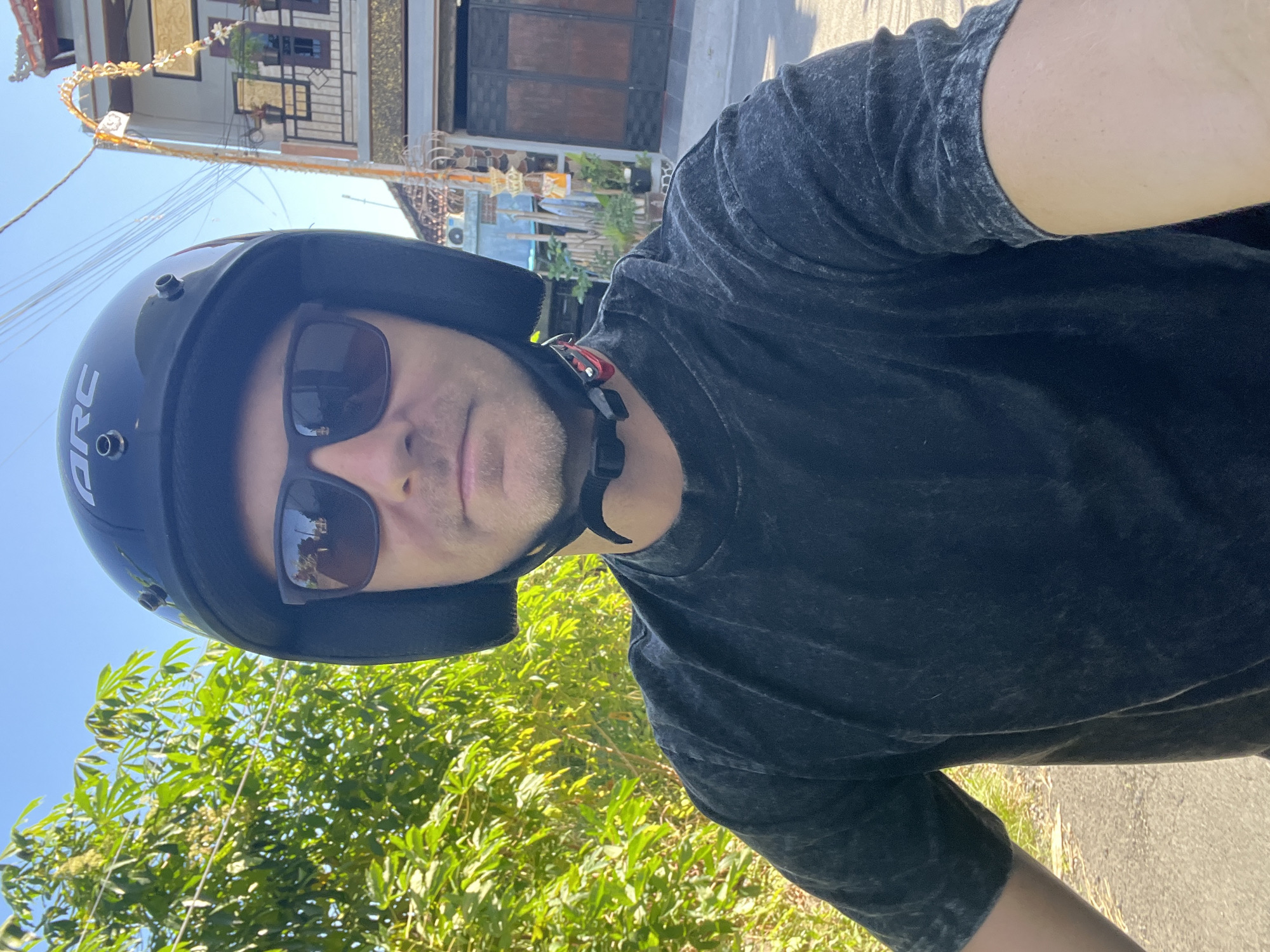A medium shot shows a person wearing a black helmet and sunglasses while riding a motorcycle. The person is wearing a dark gray t-shirt. The background is blurry but appears to be a street in Sukawati, Indonesia, with buildings and lush greenery. The sun is shining brightly, casting shadows. The image is taken from the rider's perspective.