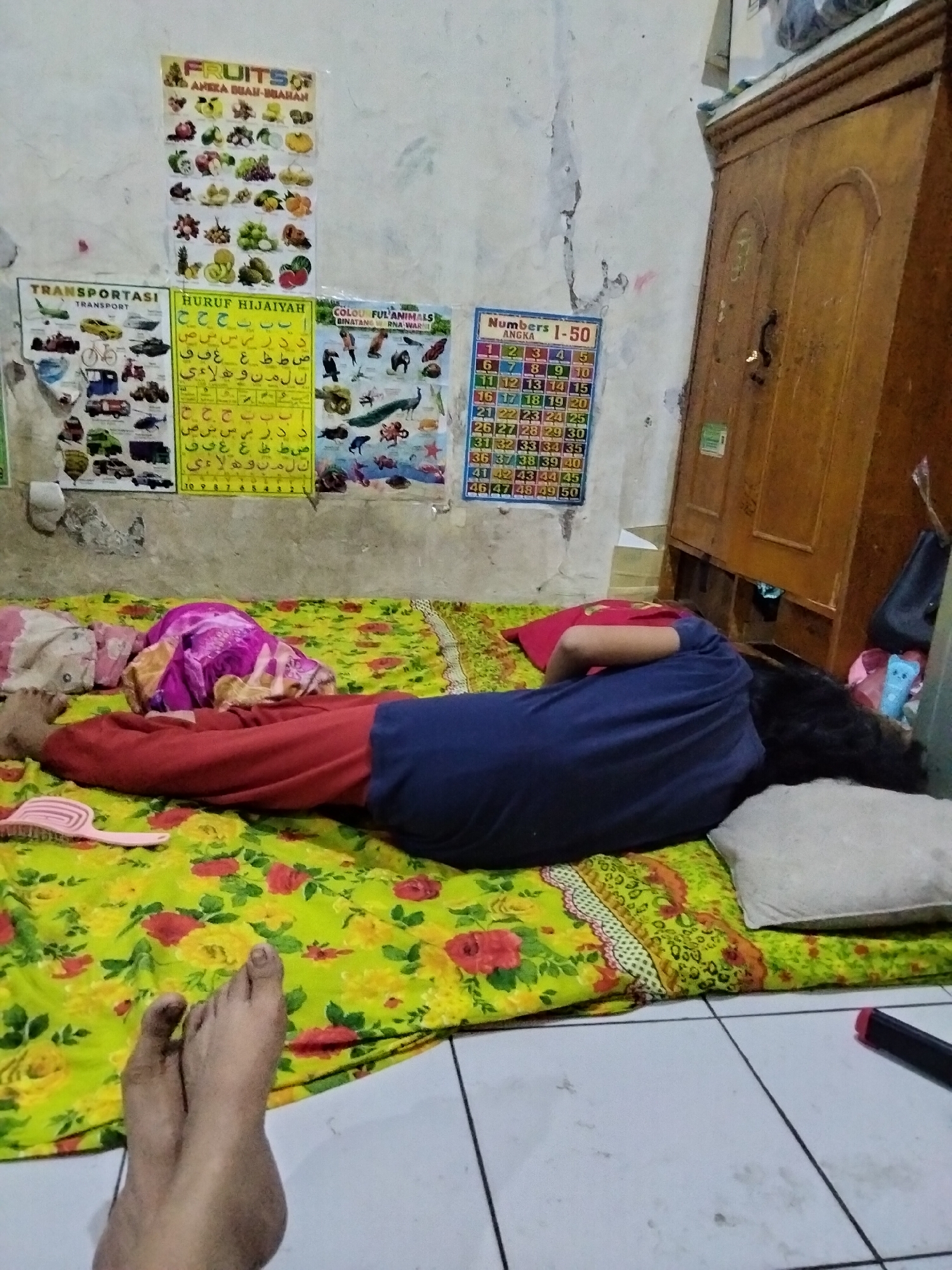 A person is lying on their stomach on a floral-patterned mat on the floor, their body is oriented horizontally across the image. The individual is wearing a blue t-shirt and red pants. Their head is turned to the right, facing away from the camera, with dark hair visible. A light-colored pillow is placed behind their head. To the left of the person's body, a pink comb lies on the mat. In the foreground, a pair of bare feet are visible, facing the camera. The background features a wall adorned with several educational posters, including one titled "FRUITS", another with Arabic script, and a chart displaying numbers 1-50. A wooden wardrobe is positioned to the right of the wall. The floor is tiled. The setting appears to be a room in a house in Cariu, Indonesia.