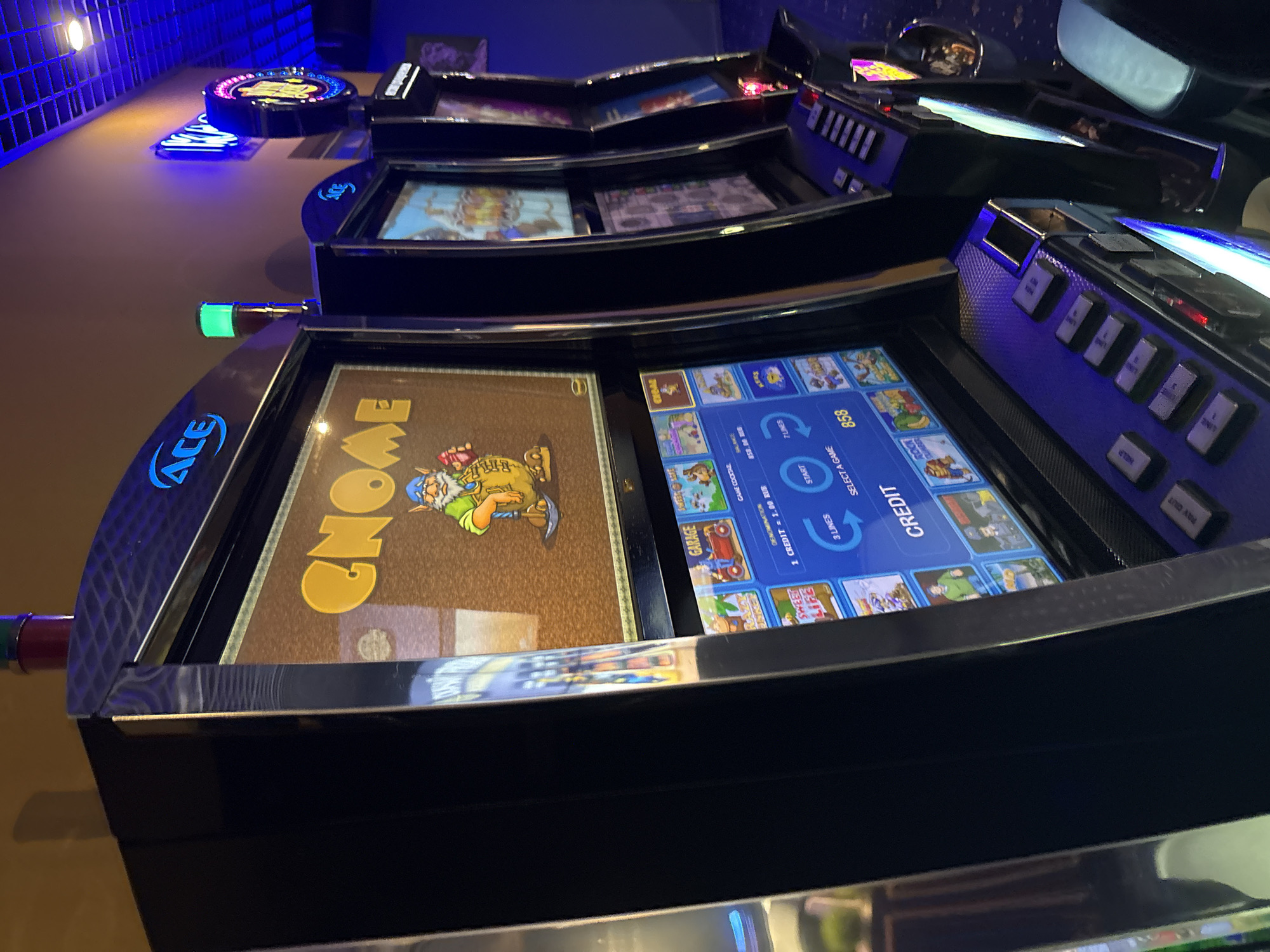 The image shows a row of slot machines in what appears to be a casino or gaming establishment. The machines are black with metallic trim and feature large screens displaying colorful graphics. The top screen of the leftmost machine shows a cartoon character, possibly a gnome, with the word "Gnome" written in large yellow letters. The lower screen displays game options, including "Garage," "Sweet Life," and "Rock Climber," along with details about credits and lines. The machines are lit by a combination of overhead lighting and the screens themselves, with blue and purple hues prominent. Some of the machines have illuminated signage at the top, including one with the "ACE" logo. The overall environment suggests a dimly lit, modern entertainment space. There are no people visible in the image. The city of Lotnoye, Russia, is not depicted or referenced in the image.