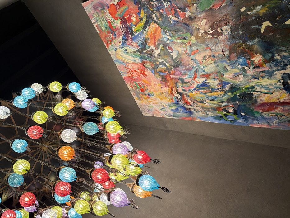 An interior scene captured from a low angle depicts a large, multi-colored chandelier and a vibrant abstract painting mounted on a wall. The location is an establishment in Budva, Montenegro.
The chandelier, positioned in the upper left quadrant and extending towards the center, is composed of numerous small, ornate glass lamps. These lamps are teardrop or onion-shaped, featuring ribbed or fluted glass in various colors including red, turquoise, yellow, orange, white, and purple. Each lamp is suspended by a dark metal chain from a larger, geometrically patterned metal structure attached to a dark ceiling. Dark metal ornamentation is present at the base of each lamp. The lamps are illuminated, casting a colorful glow.
To the right, a large, rectangular abstract painting is displayed vertically against a dark gray wall. The artwork features a dense application of paint, characterized by thick impasto brushstrokes and visible drips, indicating a textured surface. The color palette of the painting is expansive and chaotic, incorporating shades of red, pink, orange, yellow, green, blue, white, brown, and black. The wall on which the painting hangs and the wall visible beneath the chandelier are consistently dark gray. The ceiling above the chandelier is dark, likely black or very dark gray. No individuals, specific actions, or direct interactions are visible within the frame.