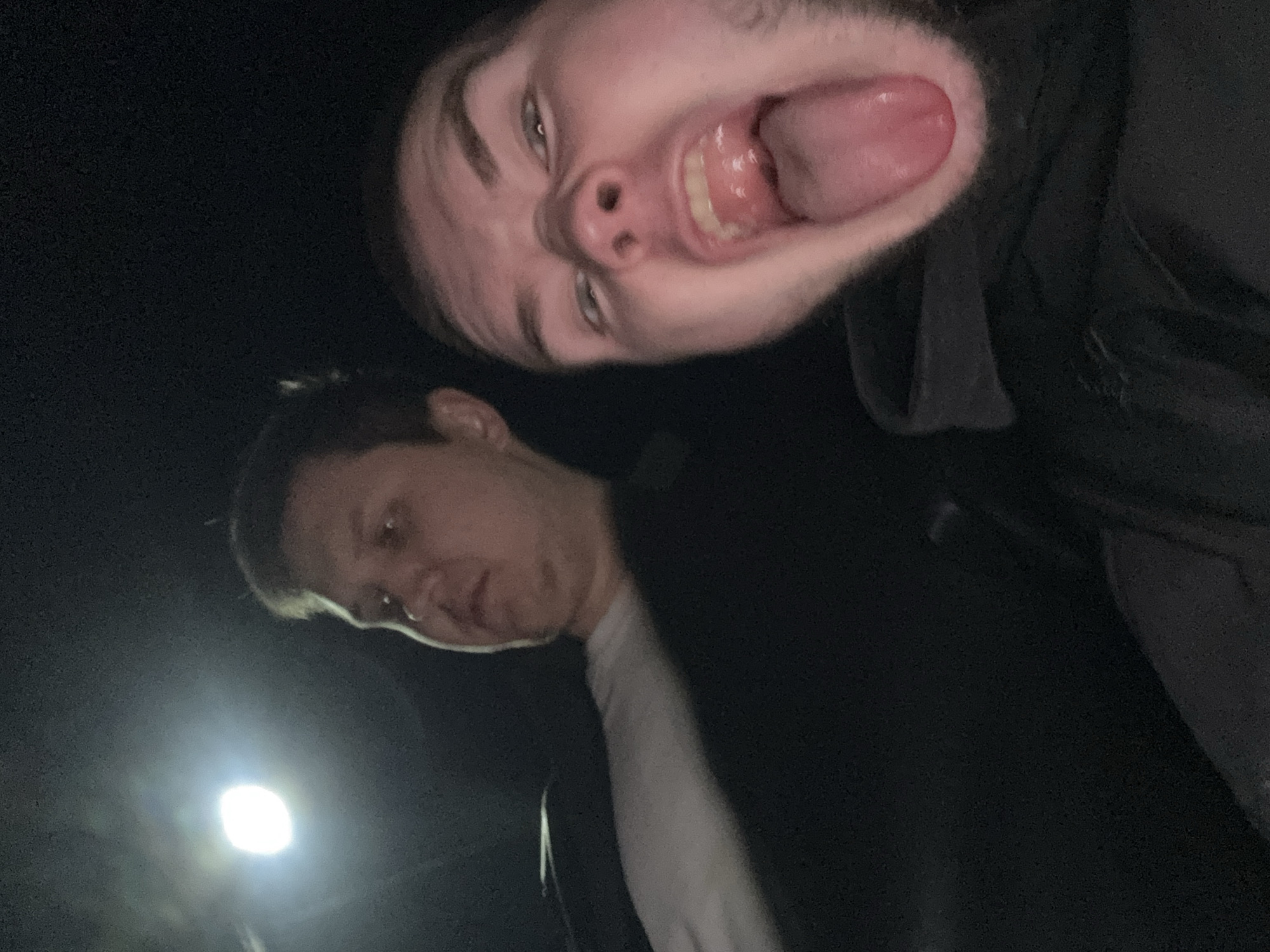 The media file features two young males in a dimly lit, dark setting, likely at night, with a prominent artificial light source visible in the lower-left background. The image is composed from a low-angle perspective, resulting in the primary subject appearing upside down.
The foreground subject is a young male with short dark hair and light-colored eyes, making a playful and exaggerated face directly towards the camera. His mouth is wide open, with his tongue extended. He is wearing a dark, possibly hooded, garment.
In the background, a second young male is positioned right-side up, looking downwards or slightly away from the camera with a more neutral expression. He also has short dark hair and is dressed in a dark jacket over a white long-sleeved shirt.
The scene captures a candid, informal moment, emphasizing the humor and playfulness of the foreground individual. The darkness of the environment suggests it is nighttime or a very dark indoor space. No specific location cues are visible within the image itself beyond the implied outdoor setting due to the street-light-like glow. There is no visible text in the image. The event is taking place in Kazan, Russia.